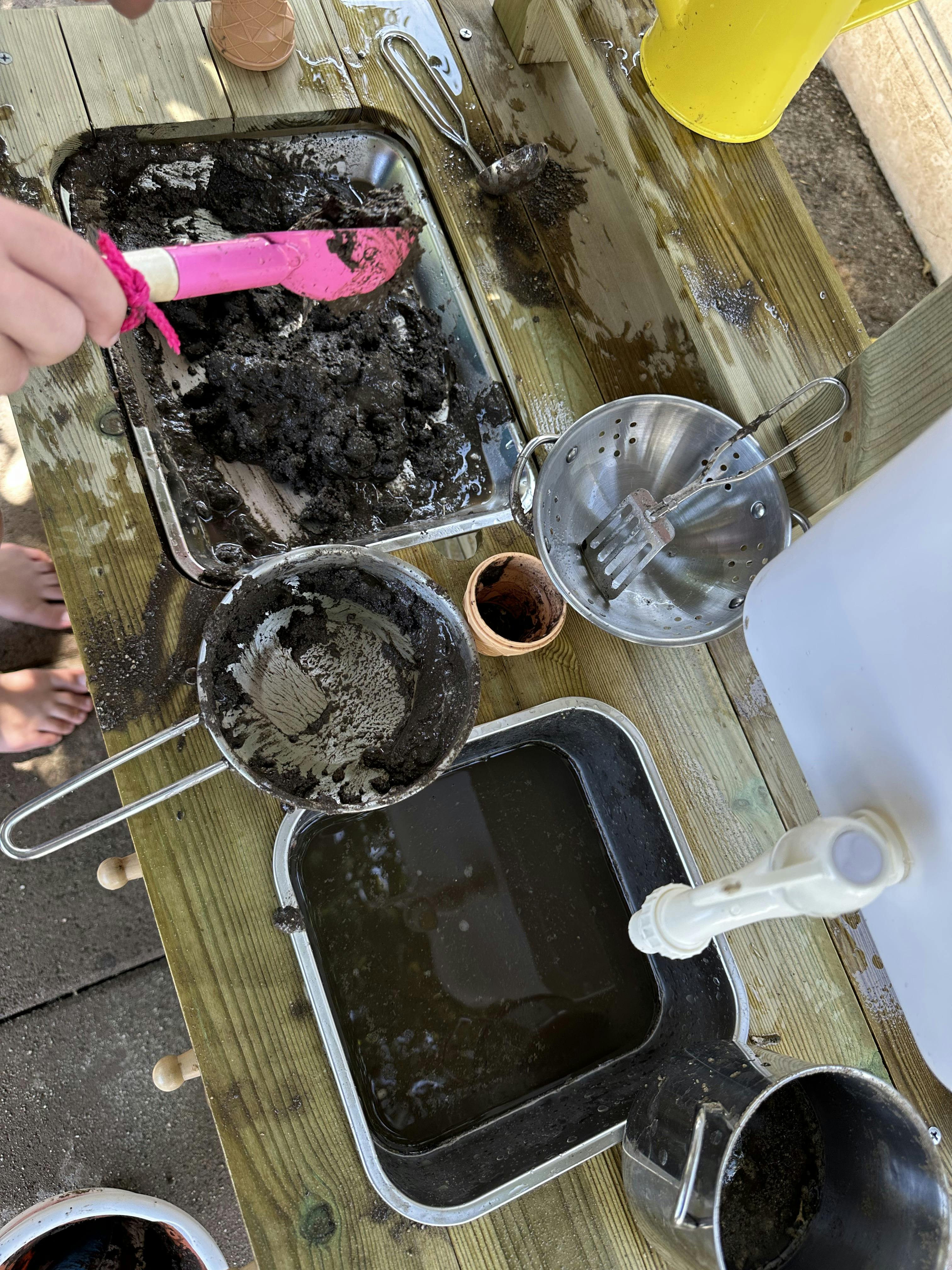 Child playing with mud and kitchen utensils on an outdoor wooden table