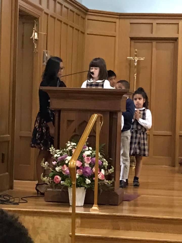 Child speaking at a podium with other children and an adult nearby