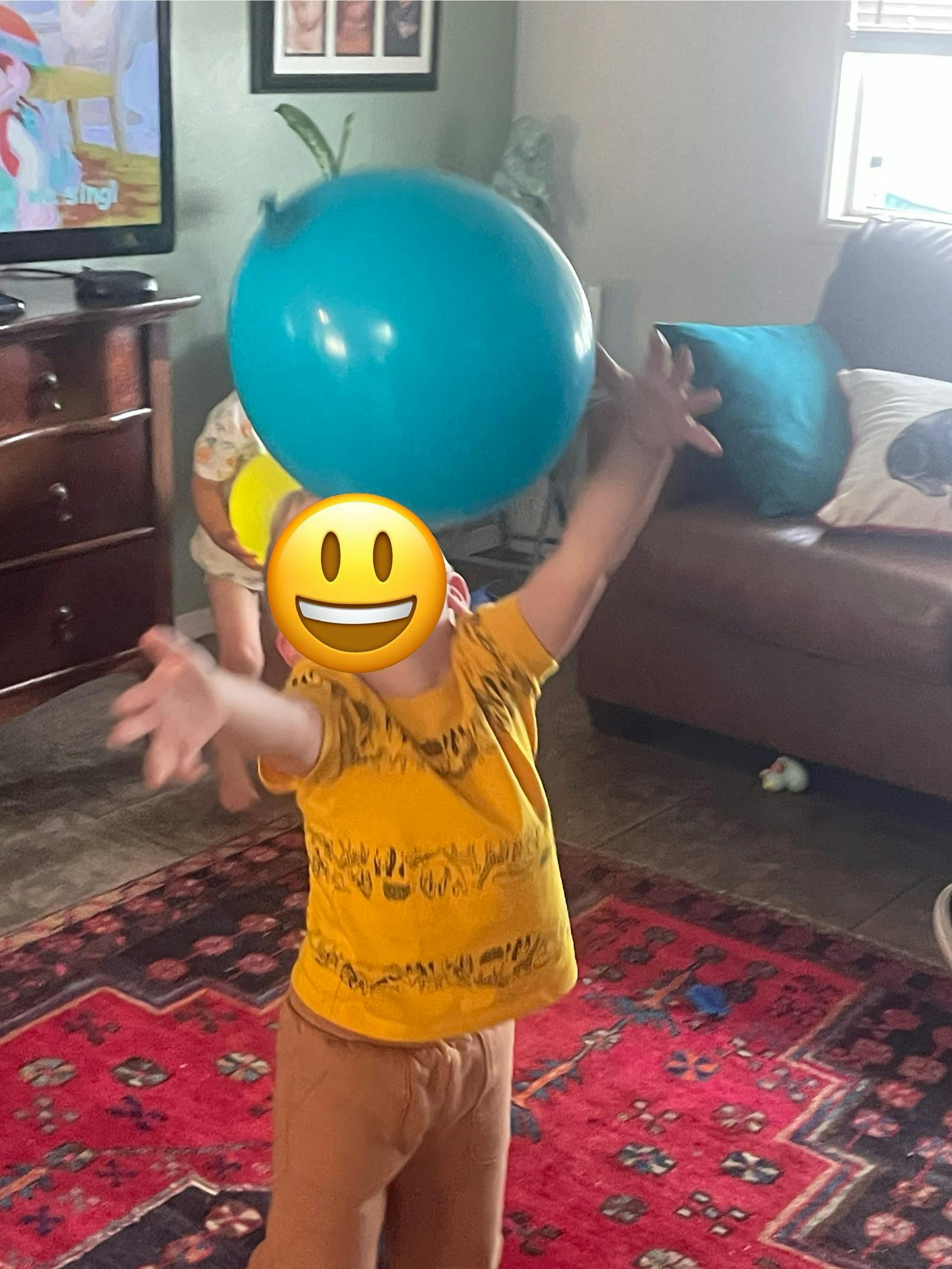 Child in a yellow shirt reaching for a blue balloon in a living room