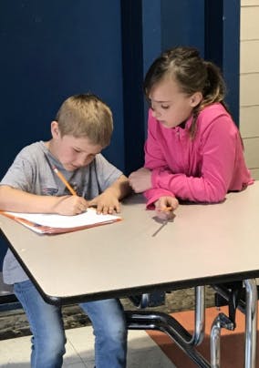Child writing at a table with another child looking on