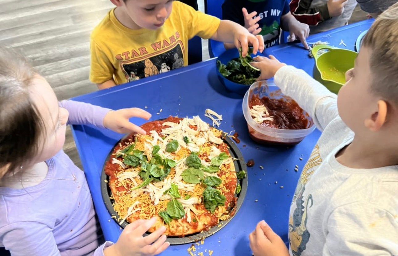 Children topping a pizza with spinach, shredded cheese, and chicken