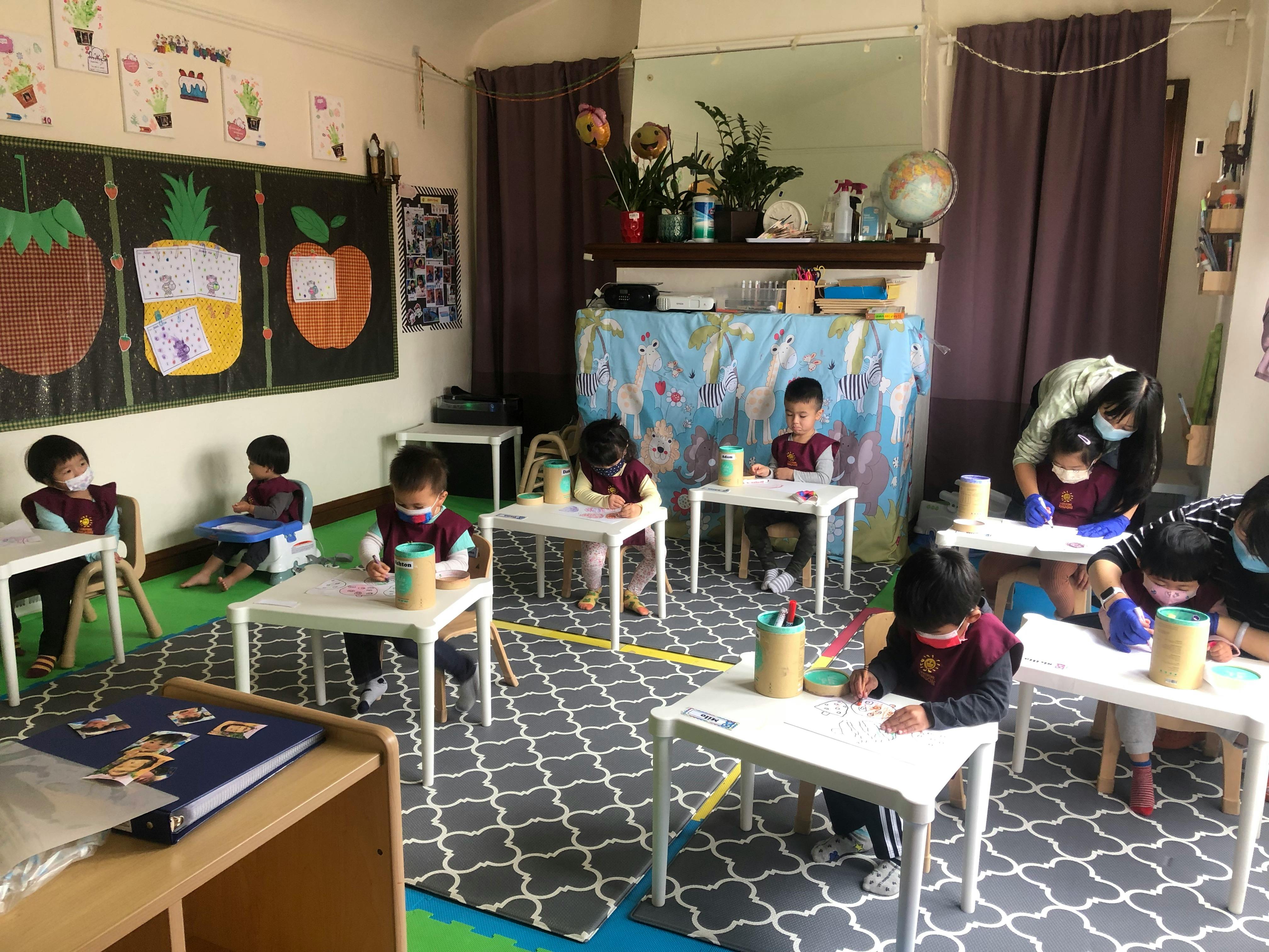 Children sitting at individual desks in a classroom, some with an adult