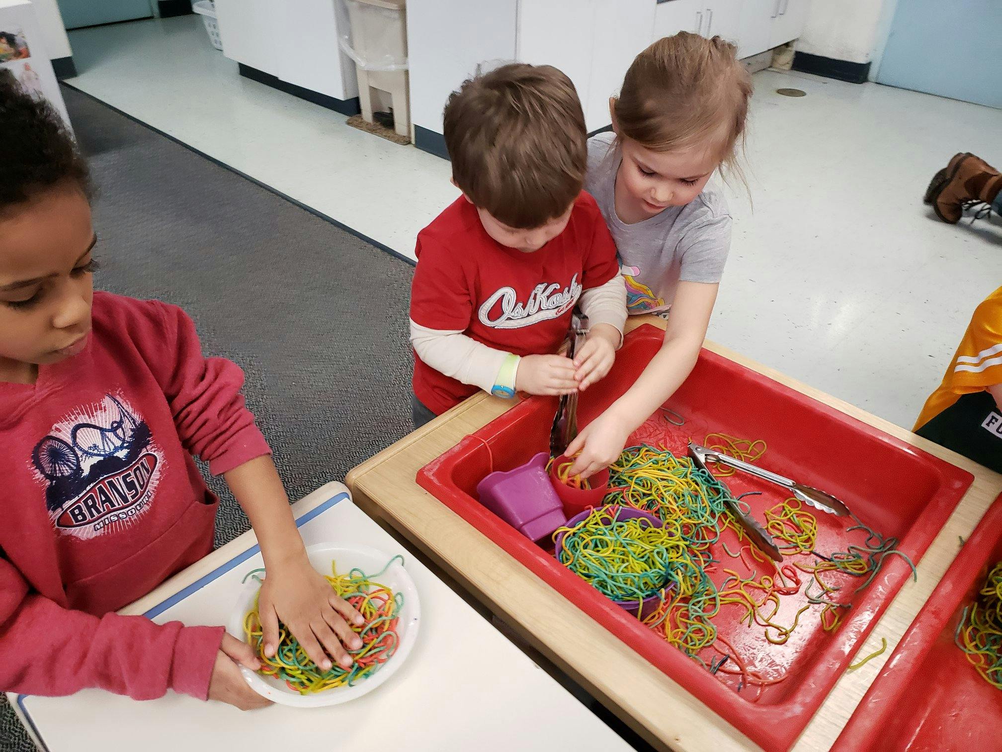 Three children playing with colorful rubber bands in a classroom