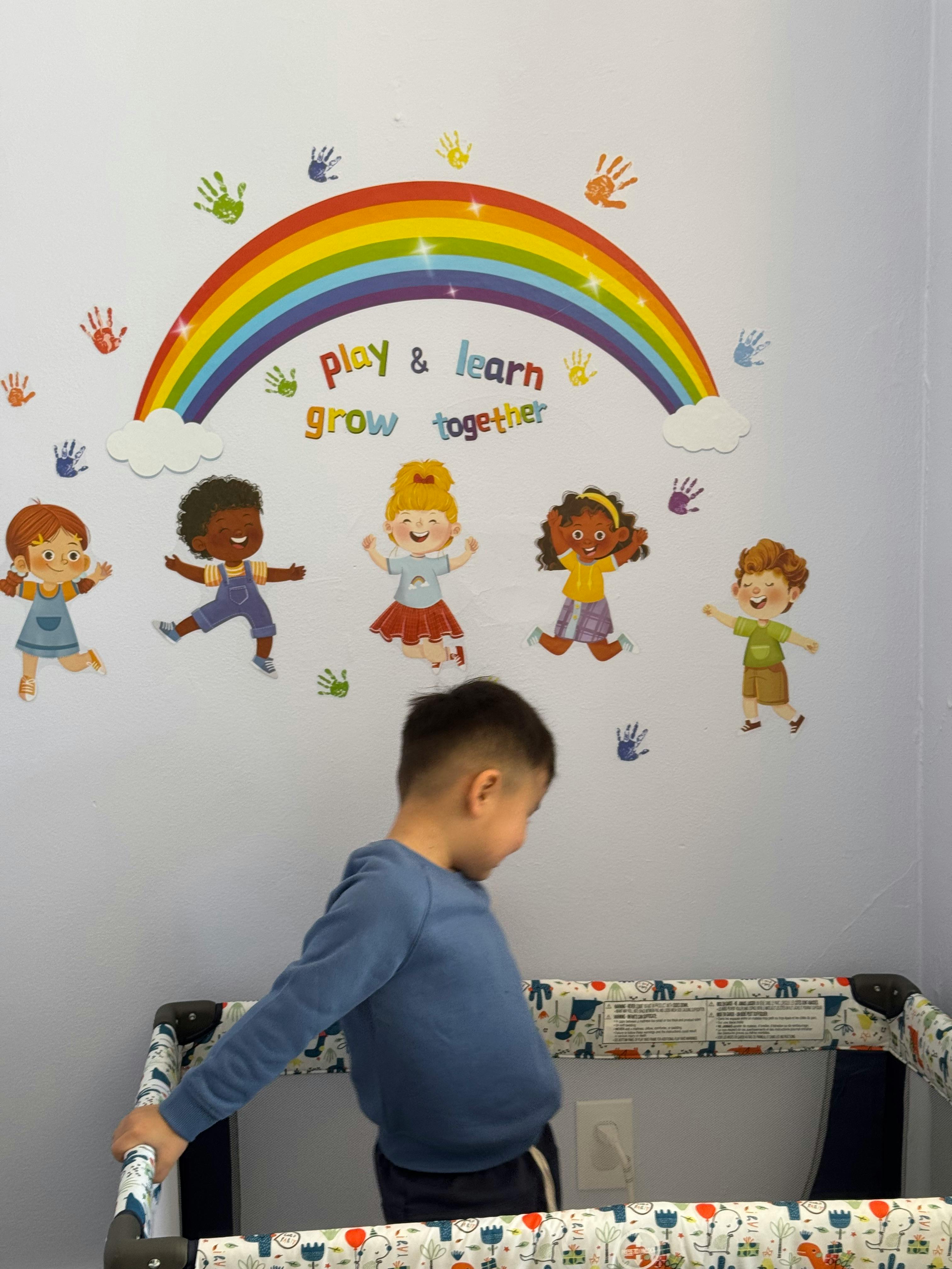 Child standing next to a playpen with a rainbow and cartoon children wall decal