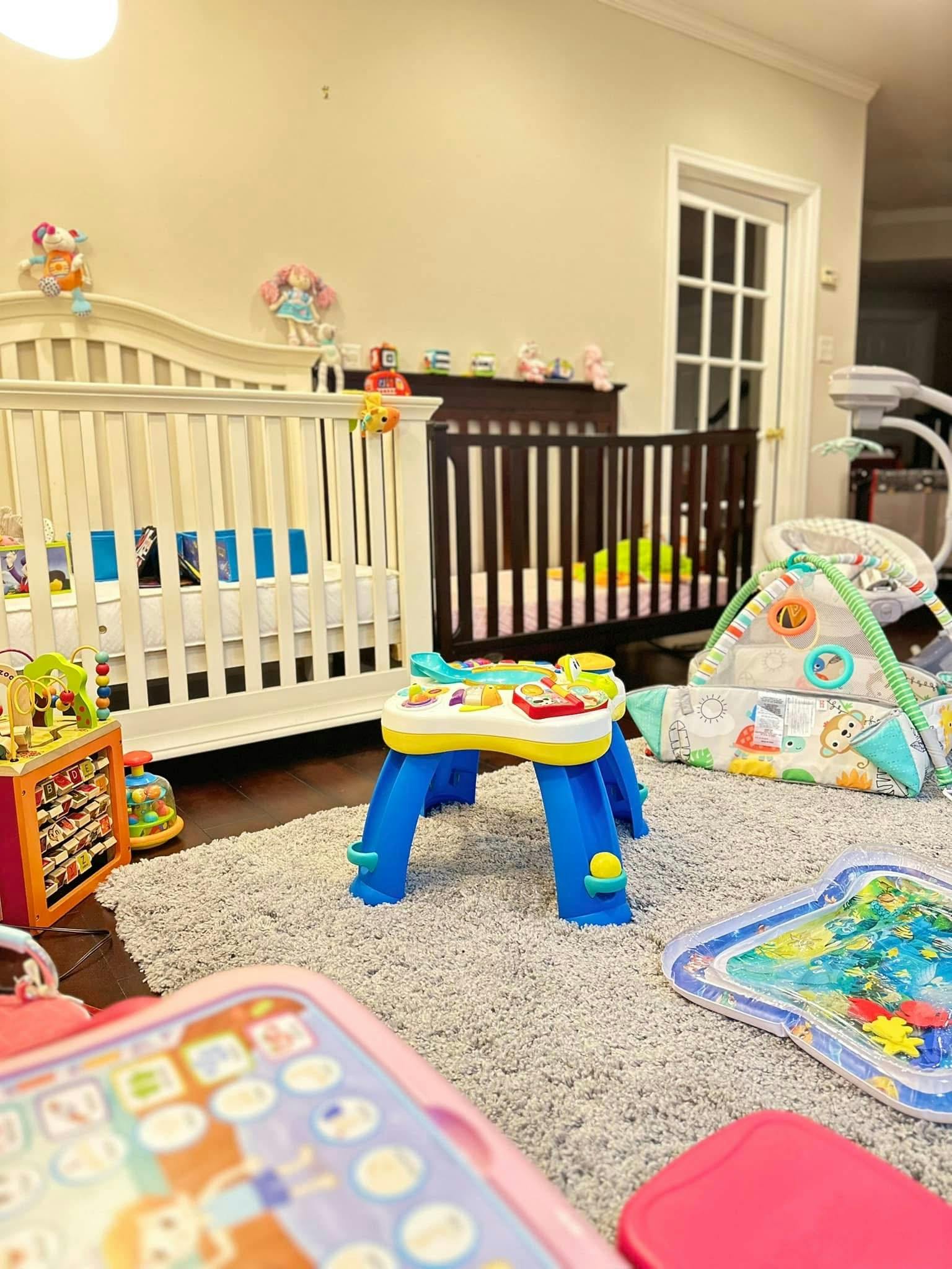 Two cribs in a room with various baby toys on a rug