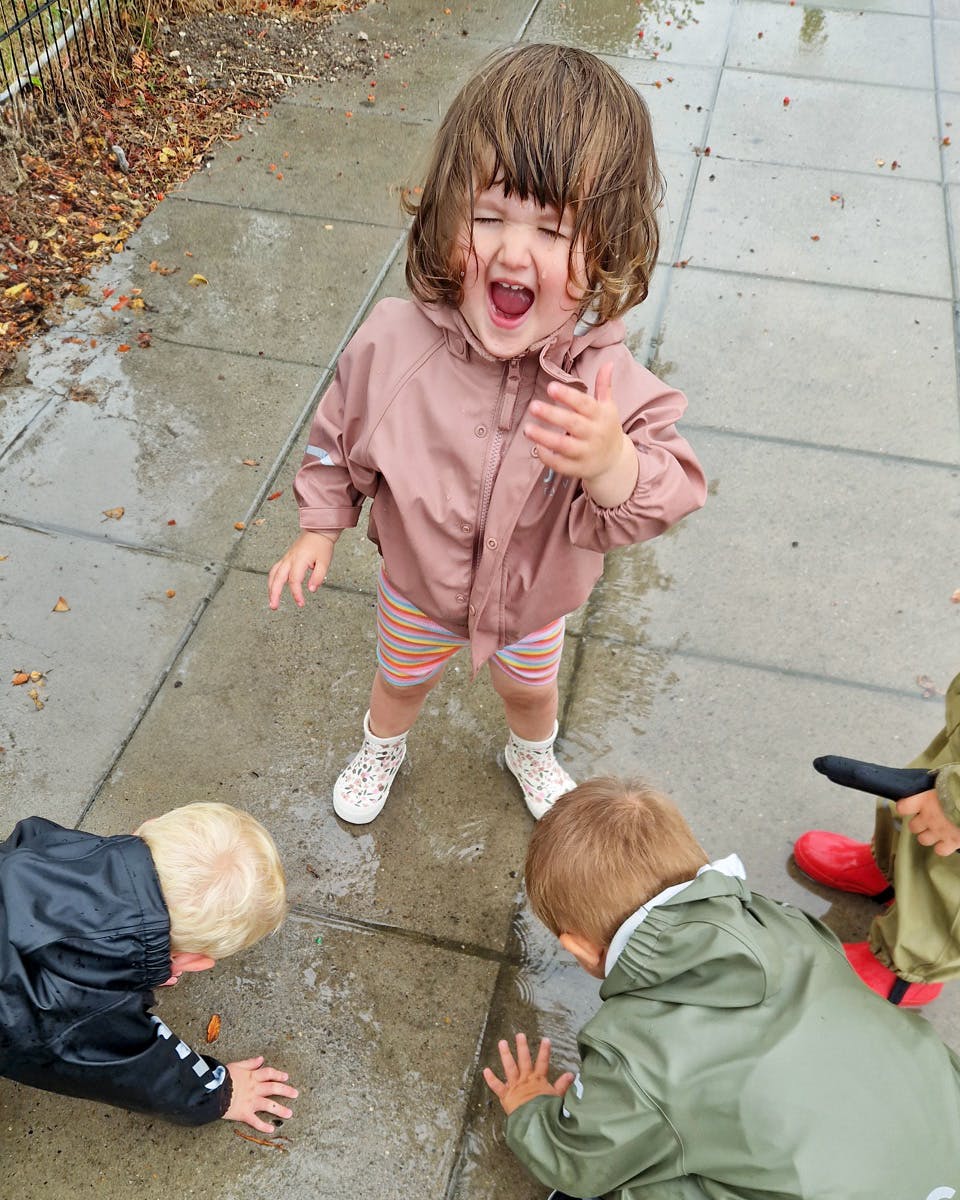 Child in a rain jacket laughing, with other children splashing in puddles