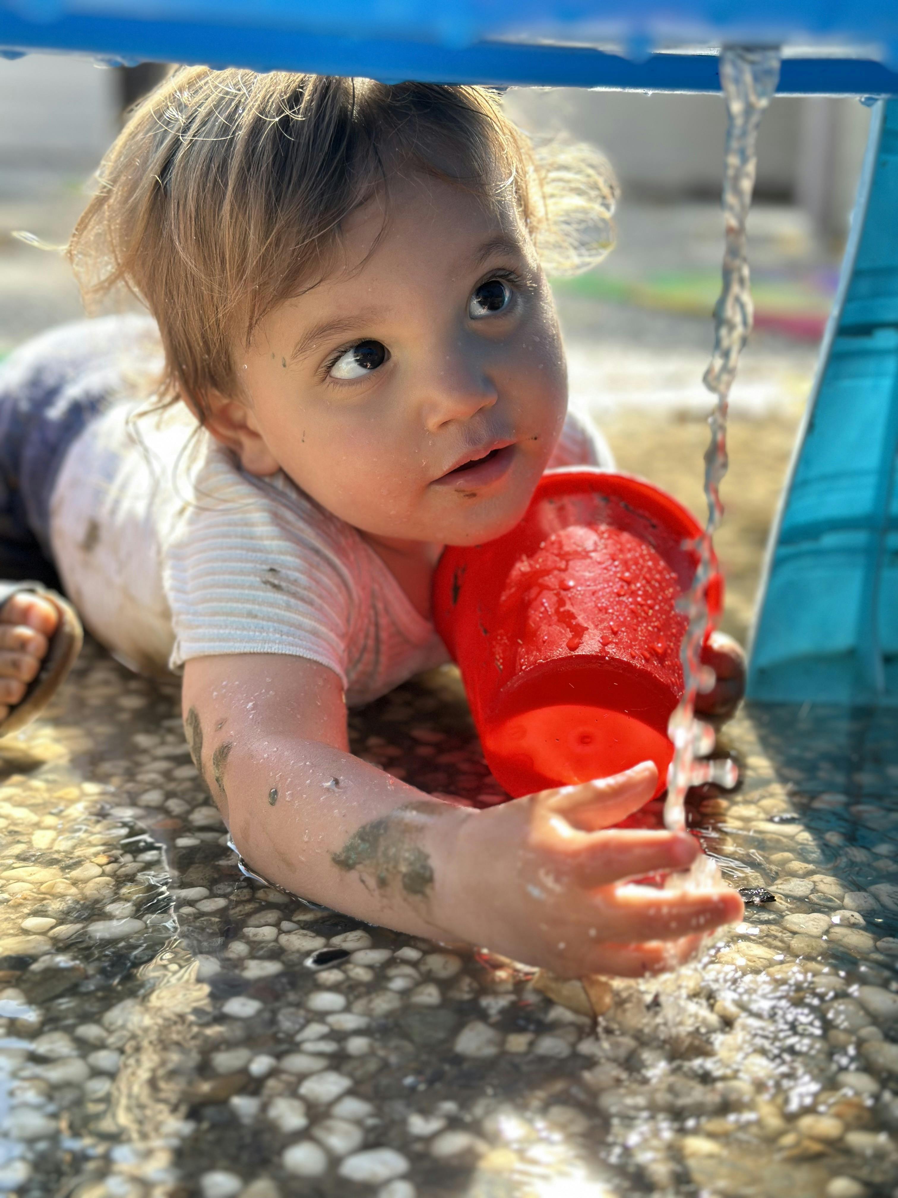 Child playing with a red cup under flowing water