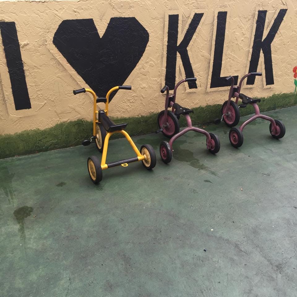 Three tricycles on a paved surface in front of a wall with "I \u2764 KLK" painted on it