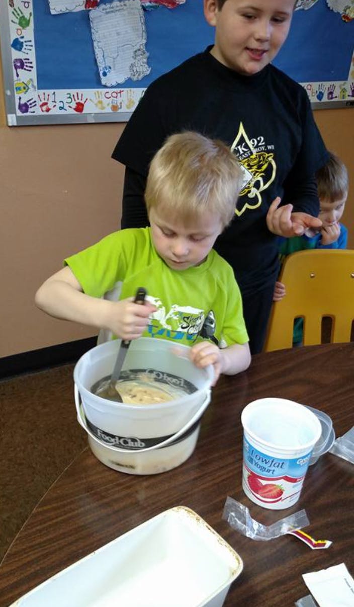 Child stirring contents in a bucket with a spoon, with another child standing behind