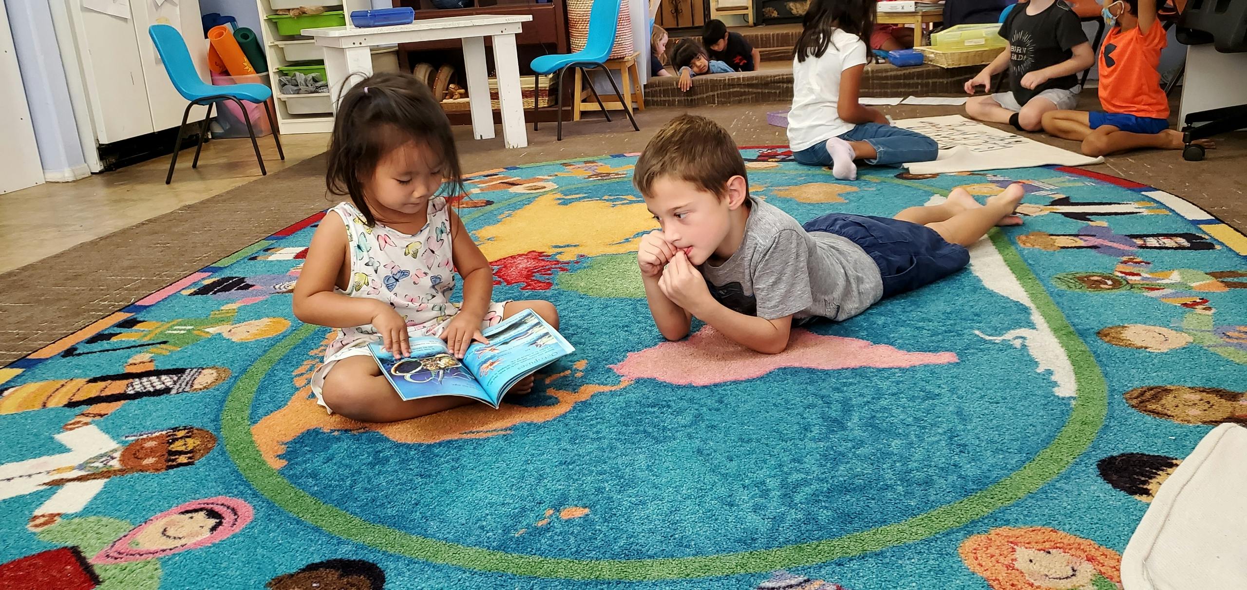Child reading a book on a rug, another child lying nearby
