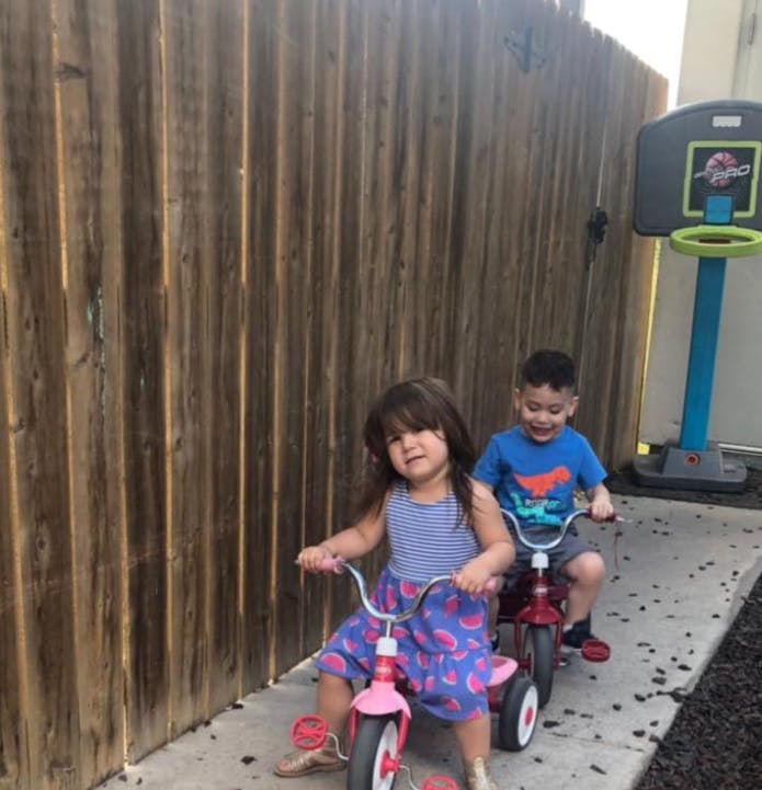 Two children riding tricycles next to a wooden fence