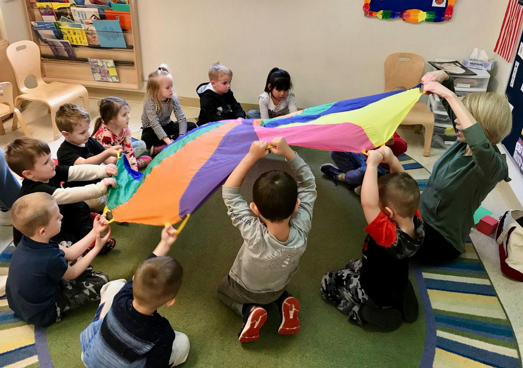 Children and an adult playing with a colorful parachute in a classroom