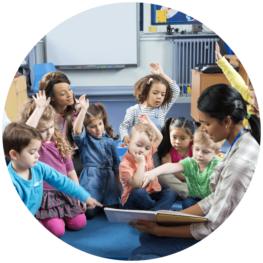 Adults reading to children in a classroom, some children raising hands