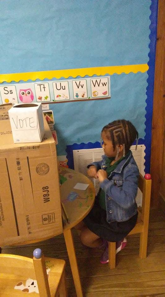 Child sitting at a small table in a classroom with a "VOTE" box