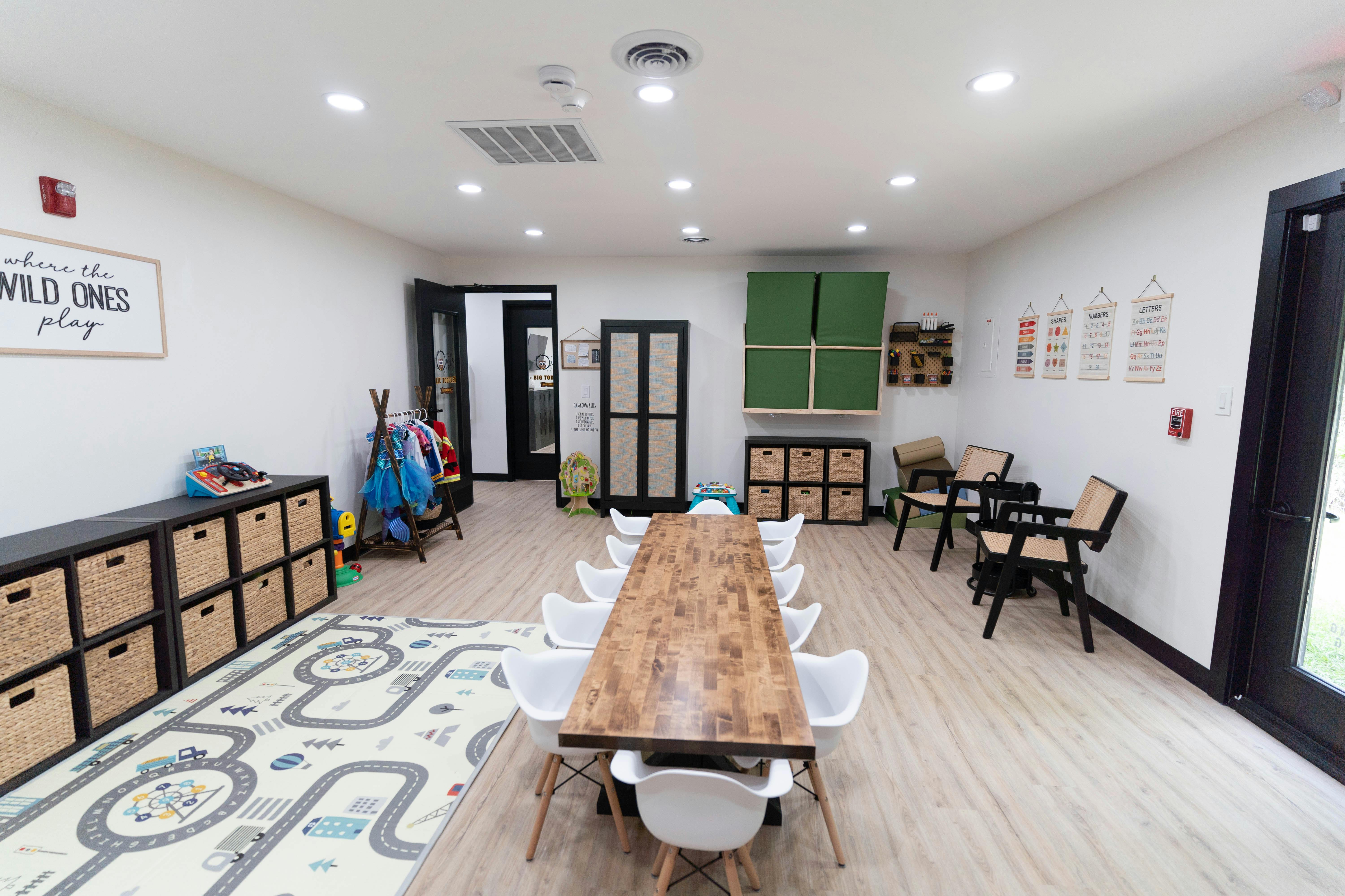 Room with a long wooden table and white chairs, storage units, and a play rug