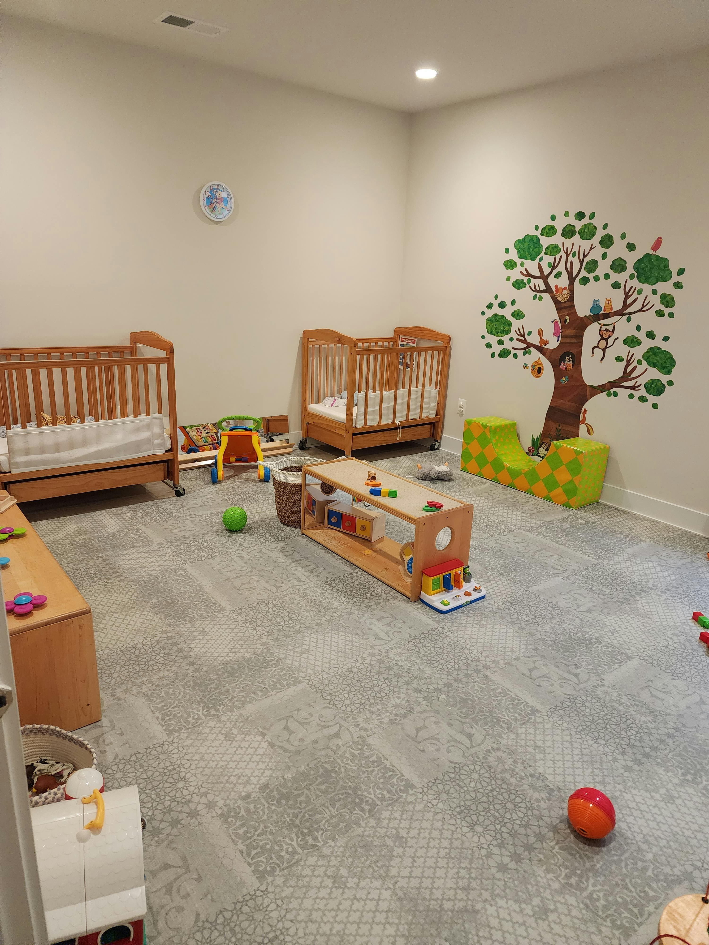 Two cribs and toys in a room with a tree mural on the wall