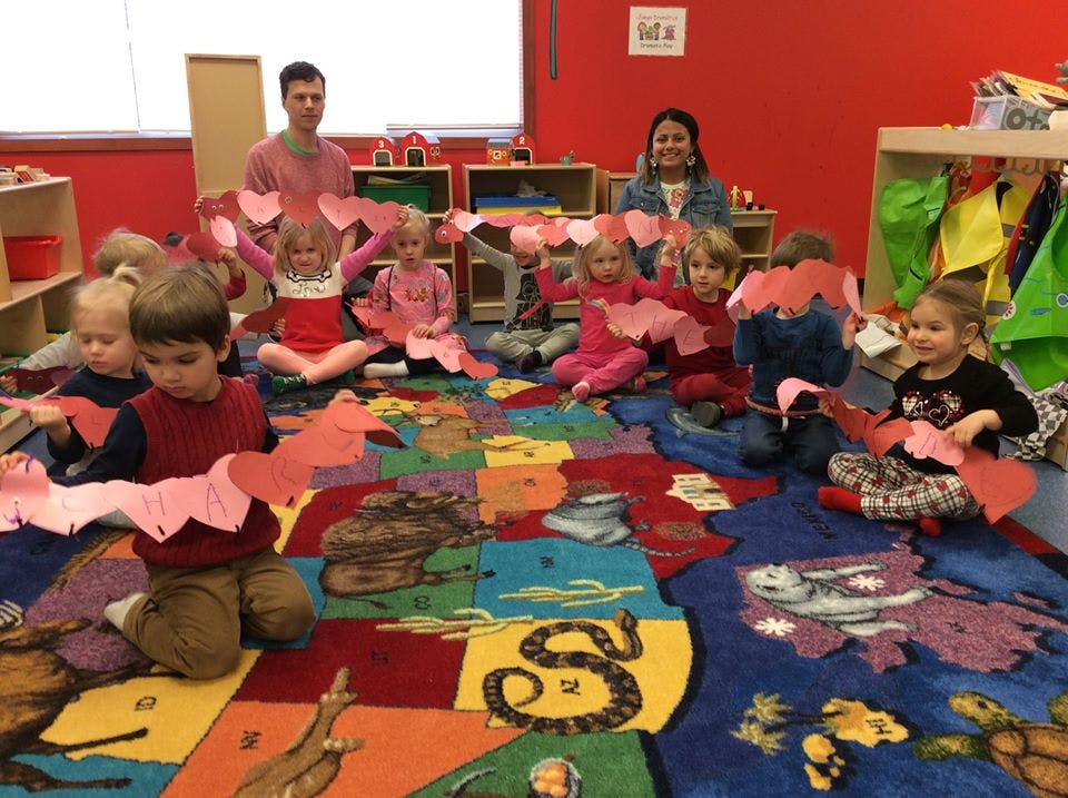 Children and adults sitting on a colorful rug holding paper heart garlands