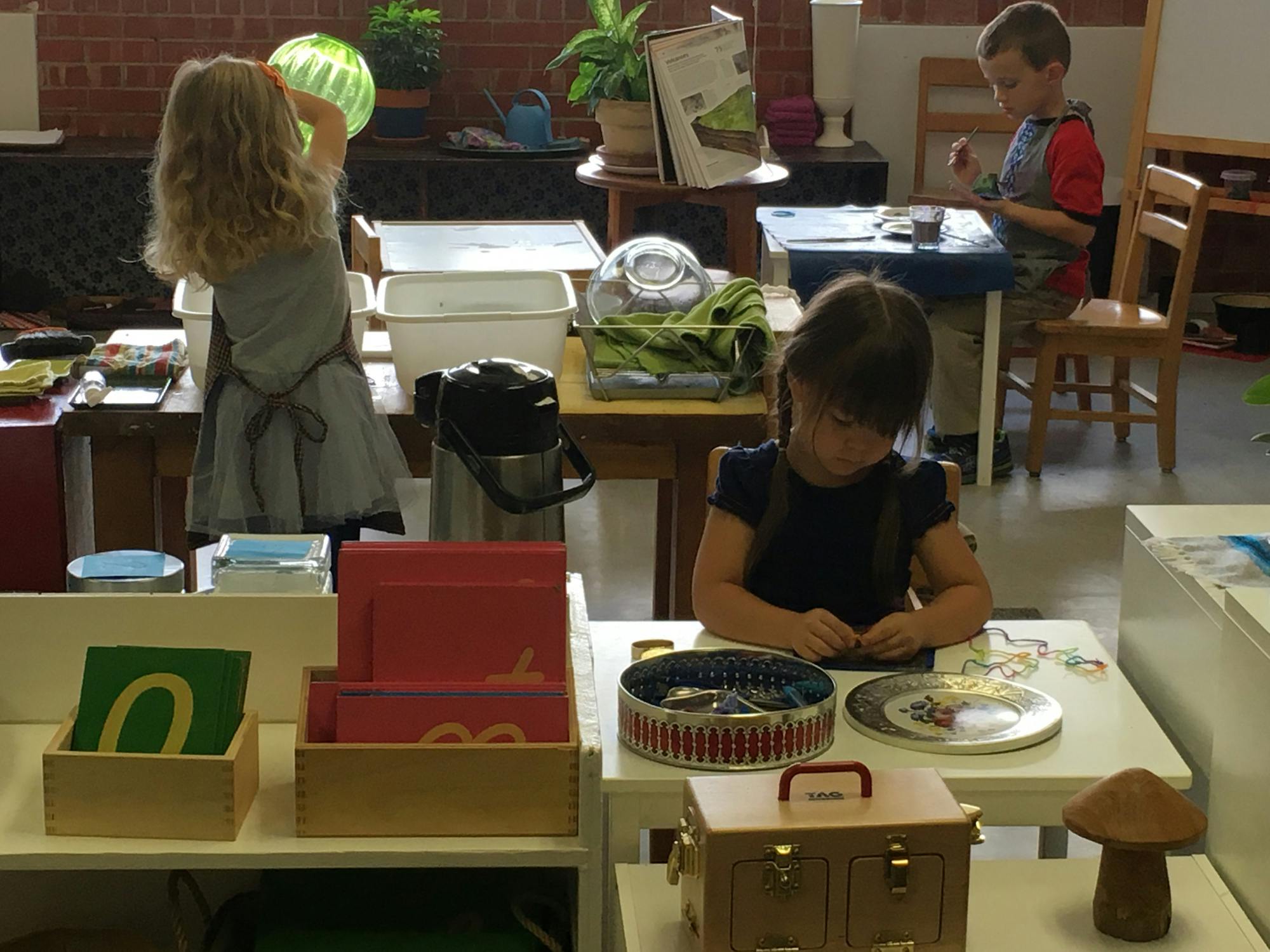 Three children in a classroom, two seated at tables, one standing