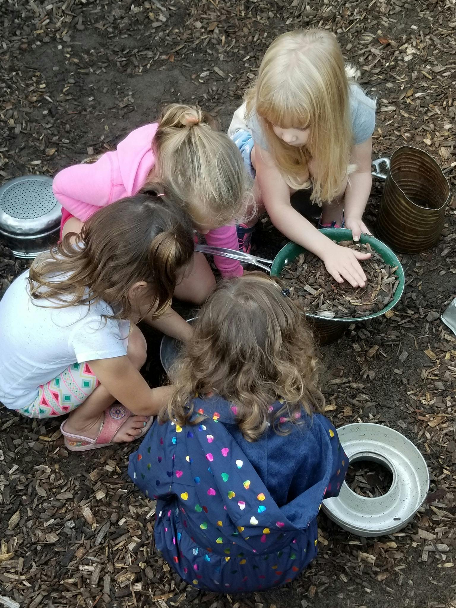 Four children playing with wood chips and pots on the ground