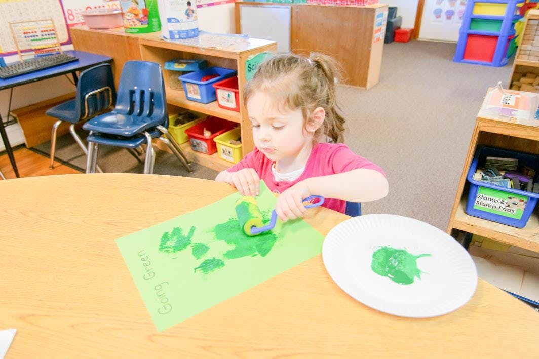 Child painting with a roller on green paper in a classroom