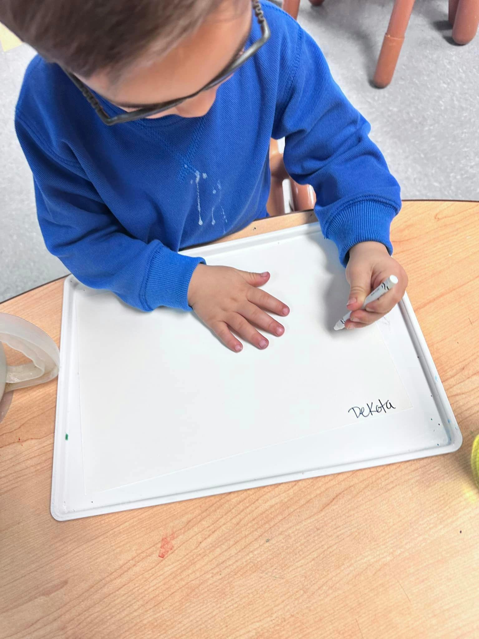 Child writing "Dekota" on a white board with a marker