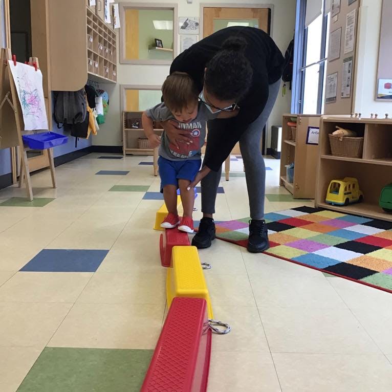 Person helping a child walk on colorful blocks in a classroom
