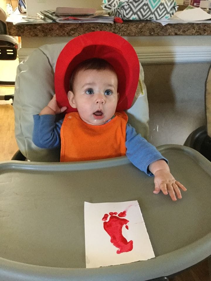 Child in a high chair wearing a red hat and orange bib, with a red footprint on paper