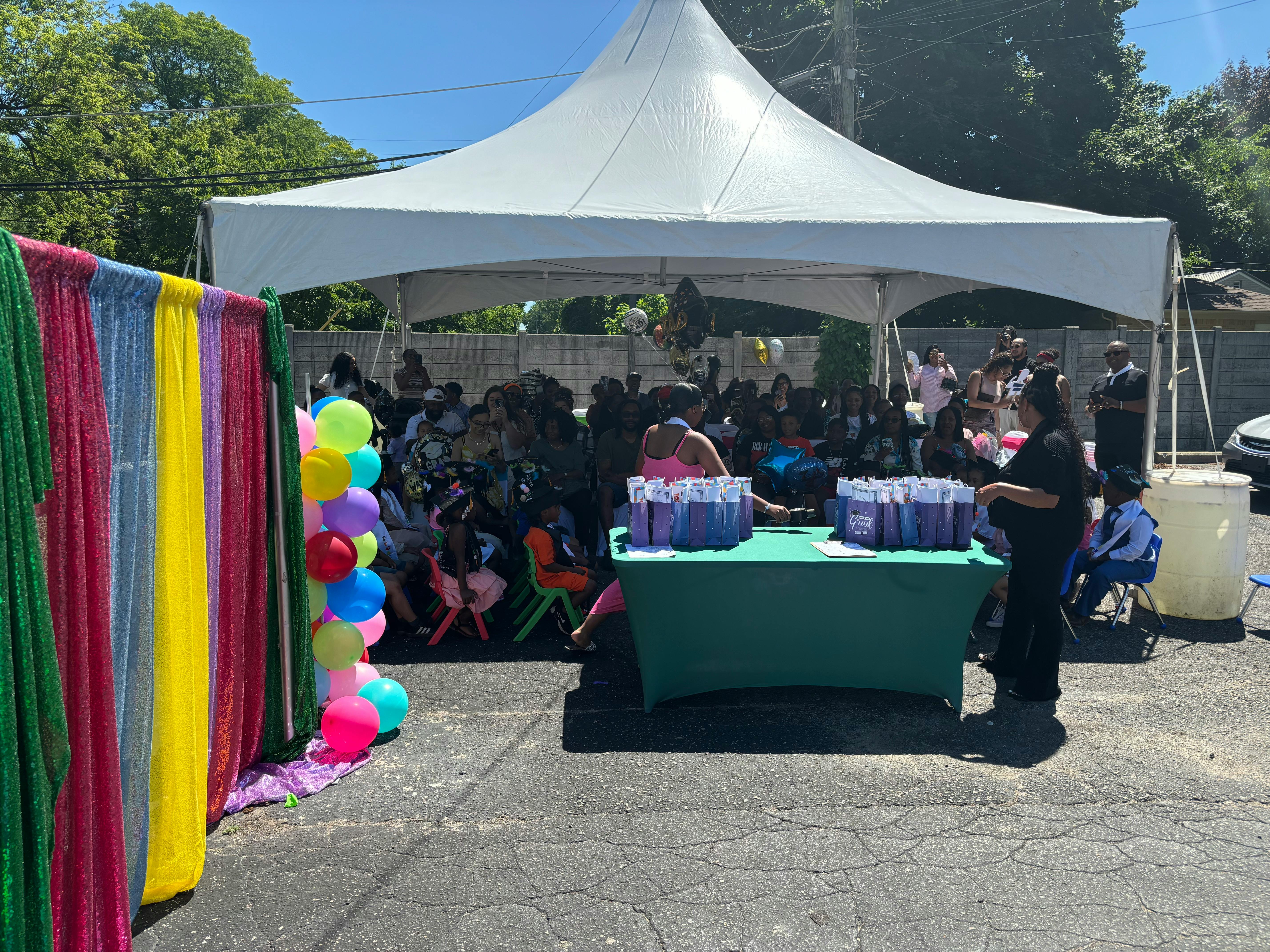 People gathered under a white tent with a table of drinks and colorful balloons