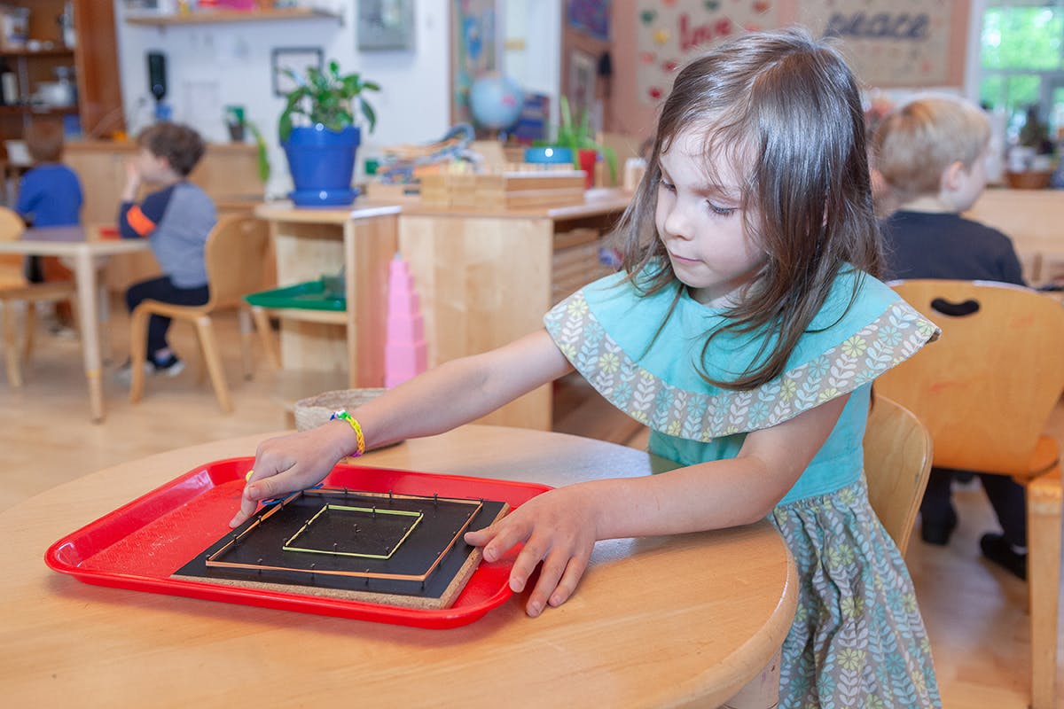 Child working with a square puzzle on a red tray at a table in a classroom