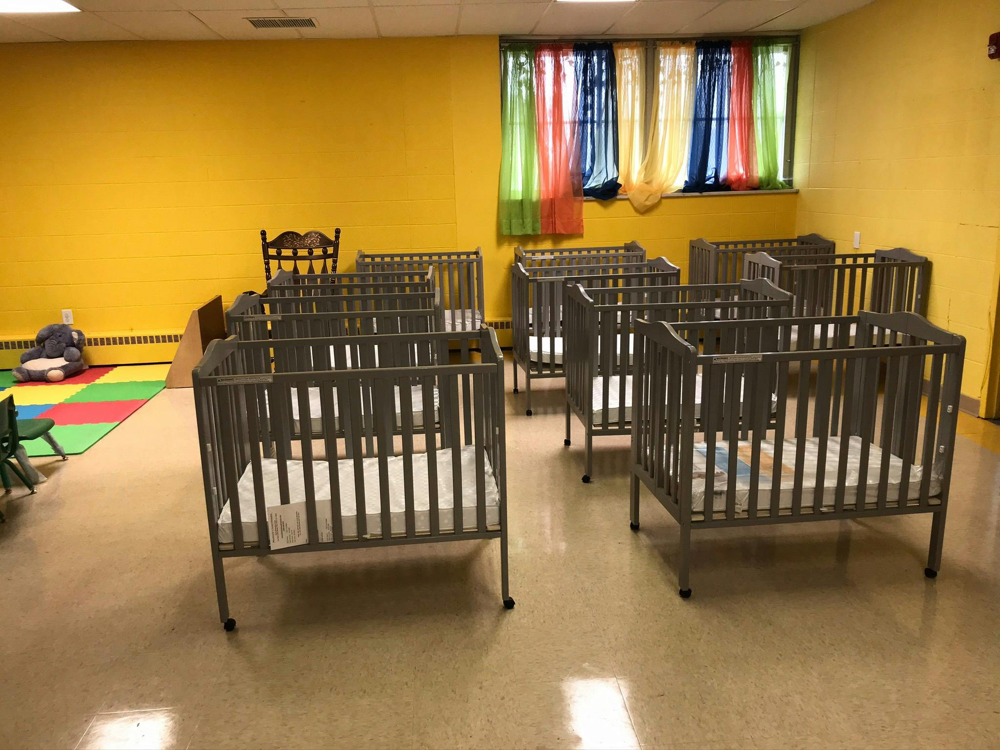 Multiple cribs in a room with yellow walls and colorful curtains