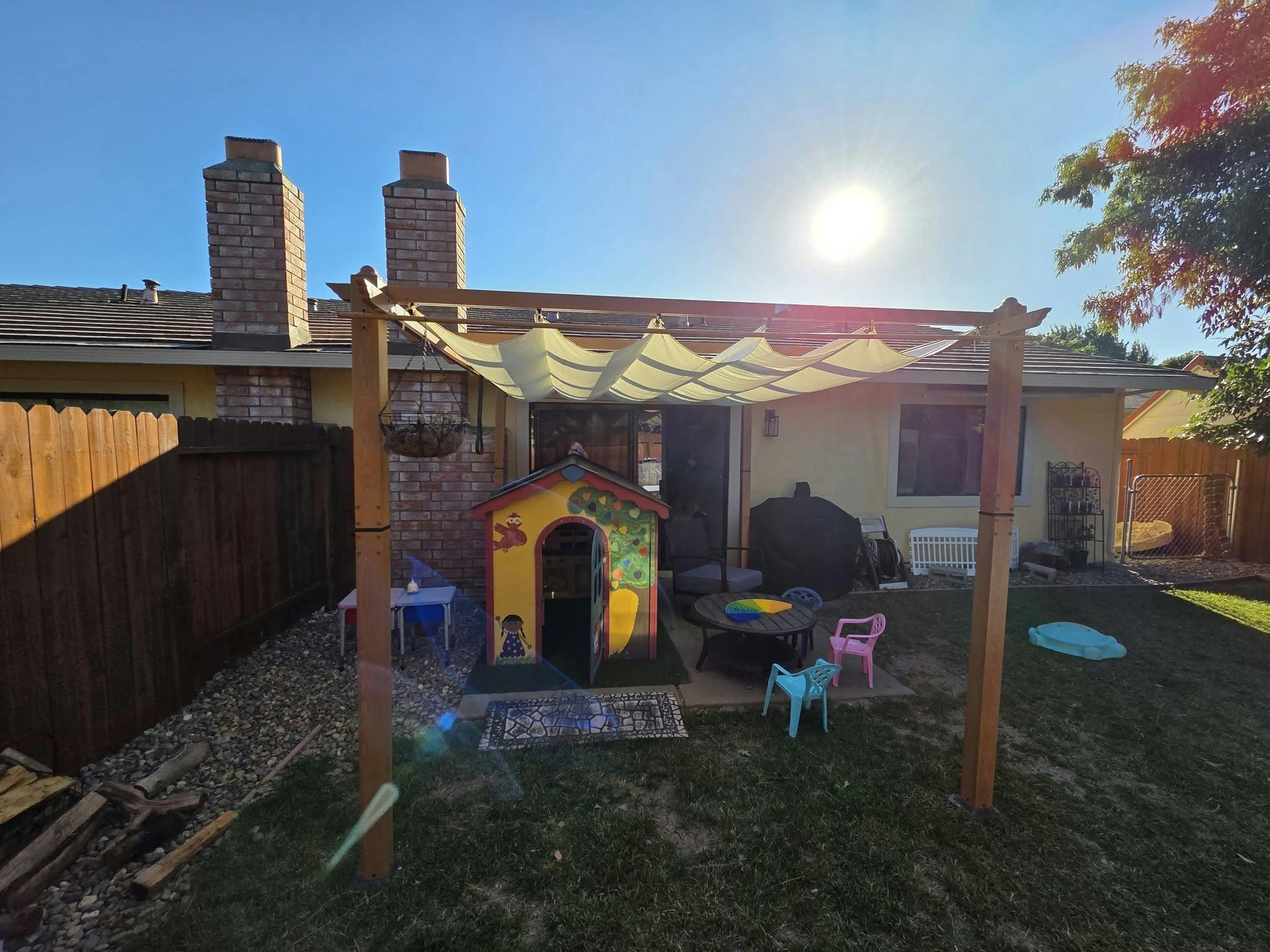 Playhouse and patio furniture under a wooden pergola in a sunny backyard