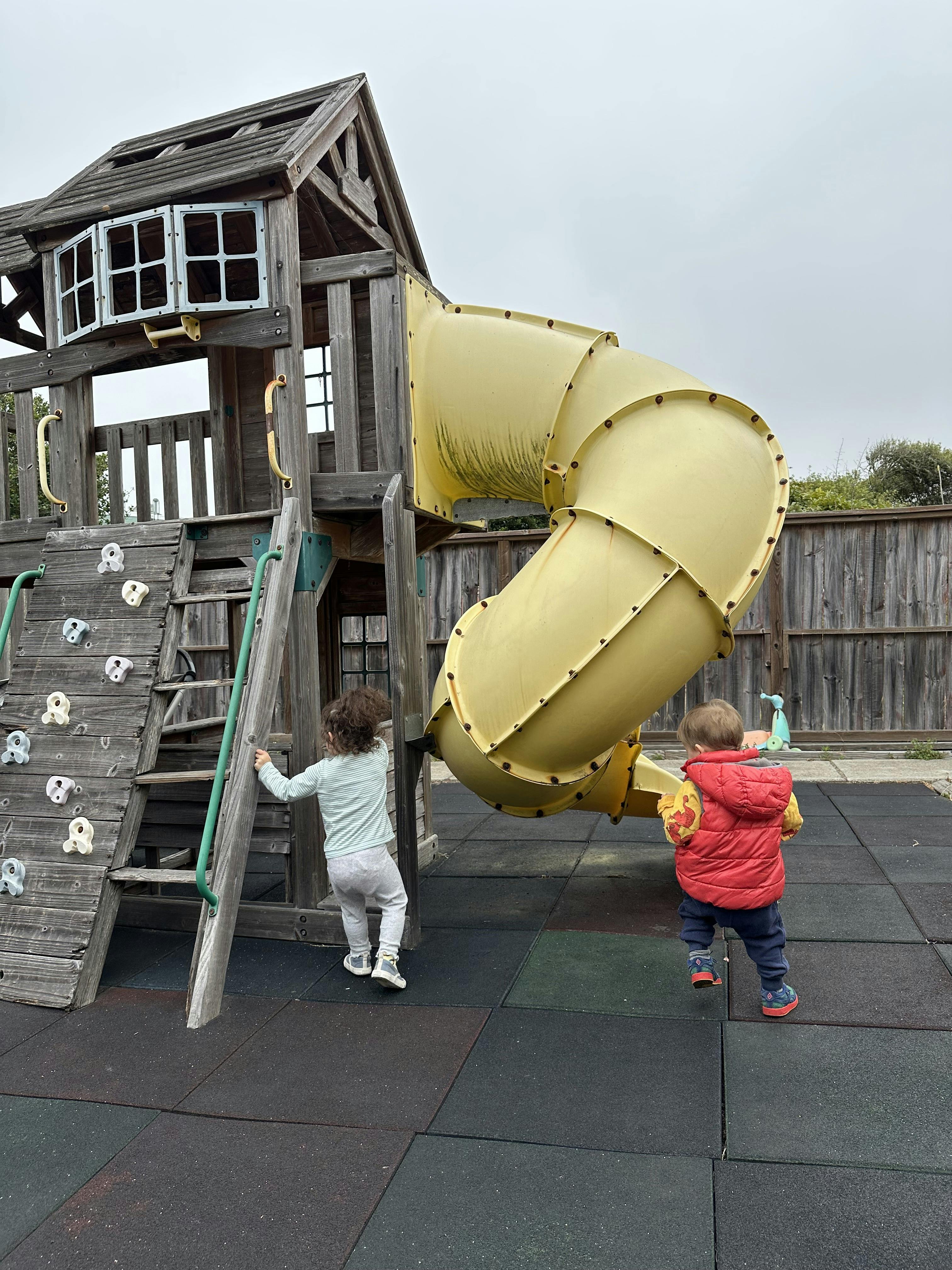 Two children on a playground, one climbing a ladder and one standing by a slide
