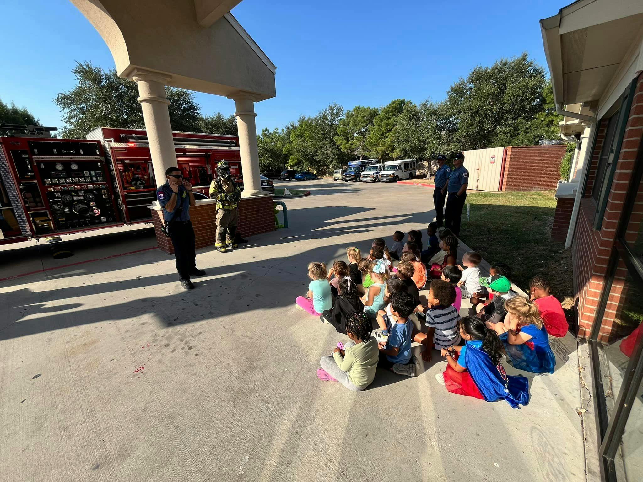 Children sitting on the ground listening to two people standing near a fire truck