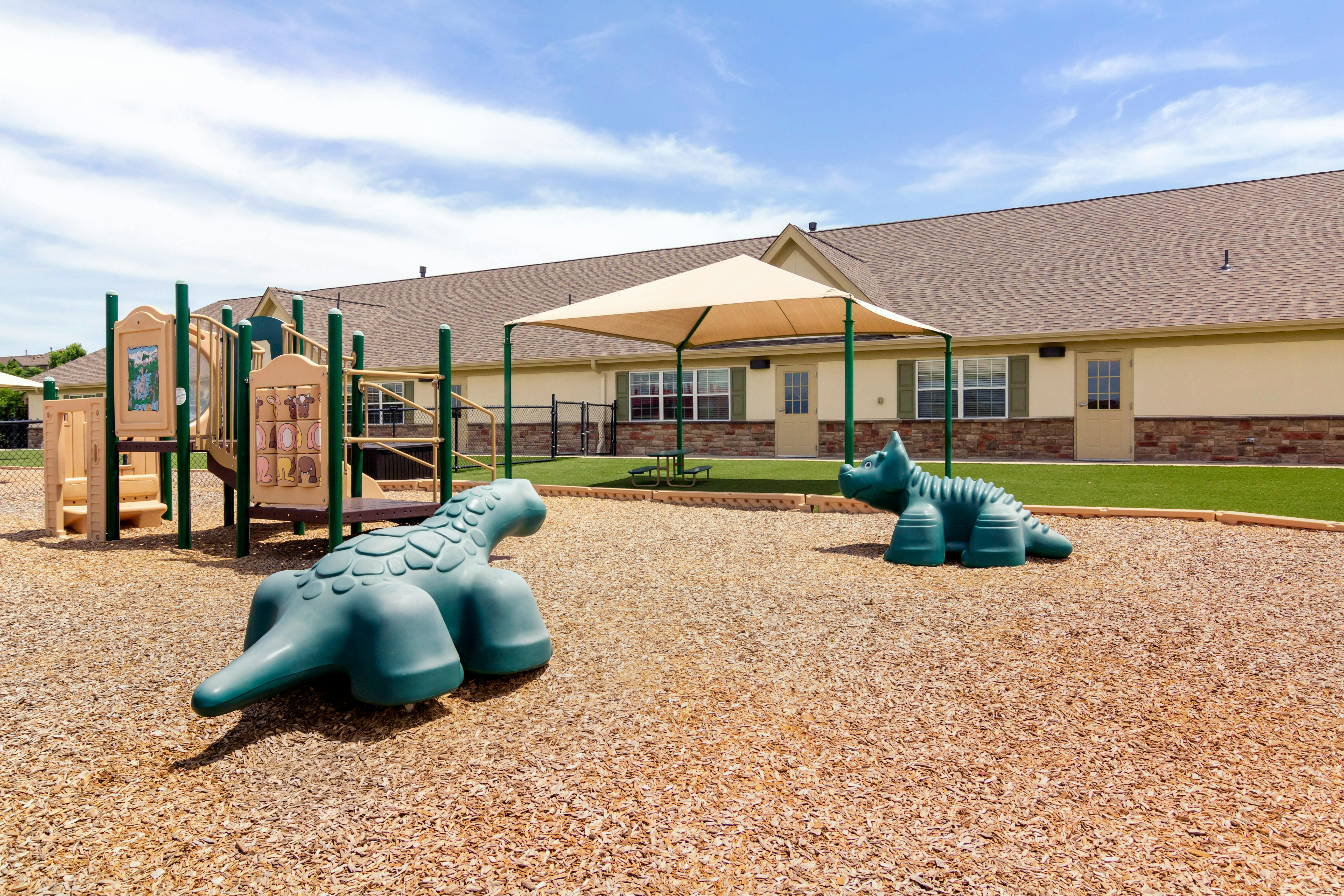 Playground with climbing structure, dinosaur ride-ons, and shade canopy over wood chips