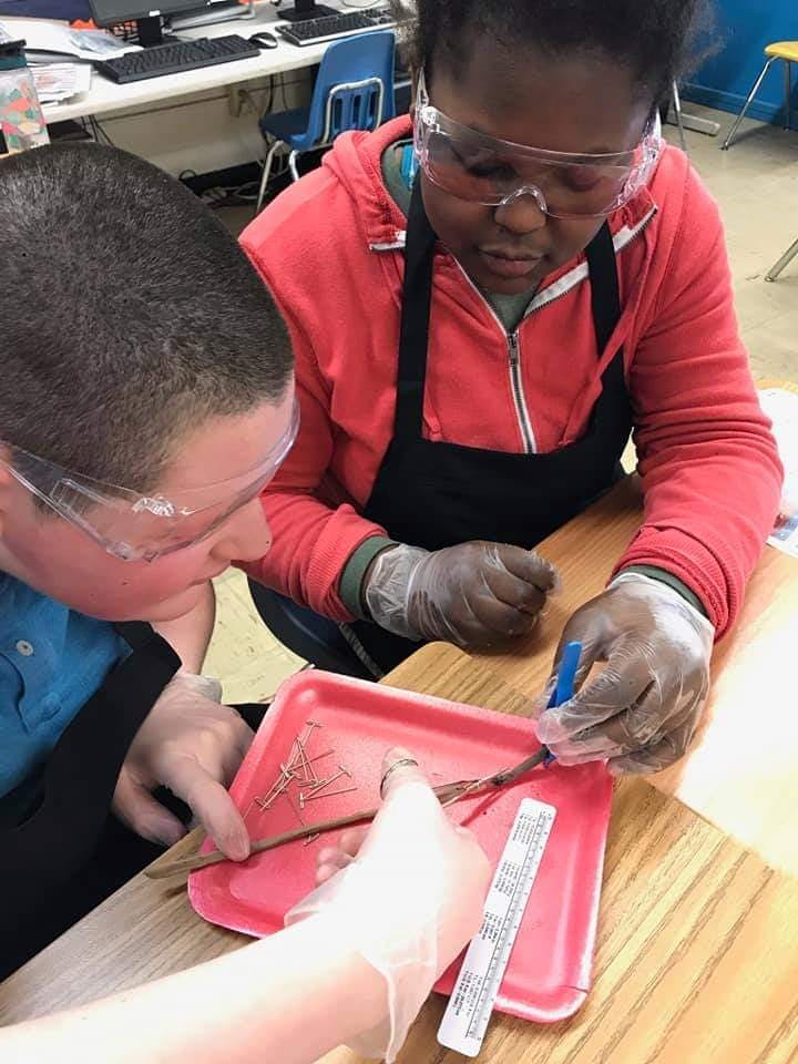 Two people in safety glasses and gloves working on a project in a classroom