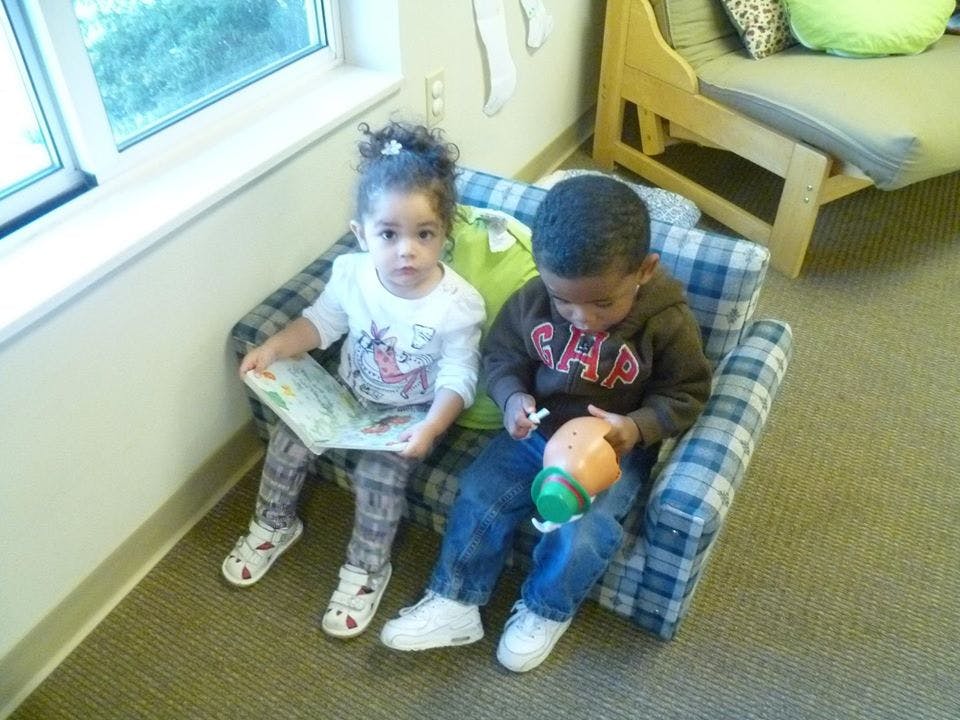 Two children sitting on a small couch, one holding a book and the other a toy