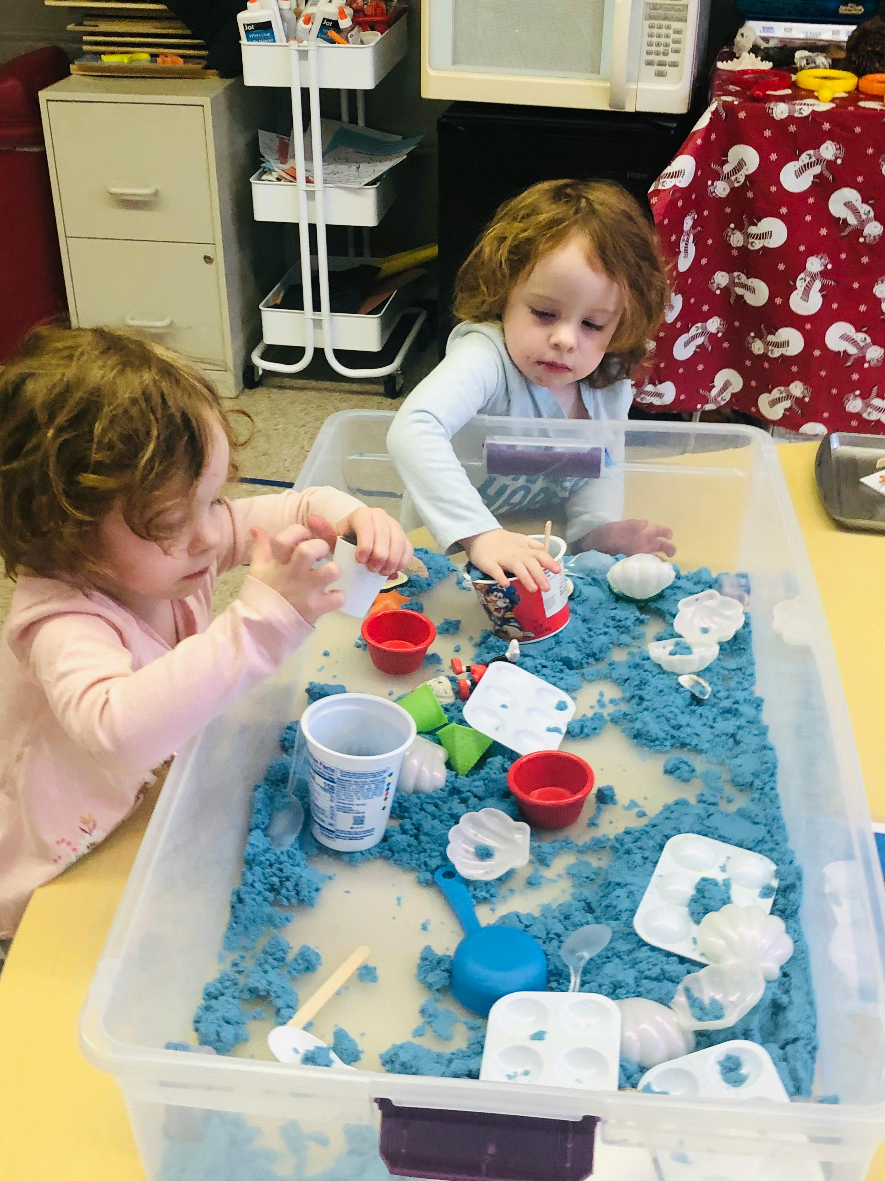 Two children playing with blue kinetic sand in a clear bin