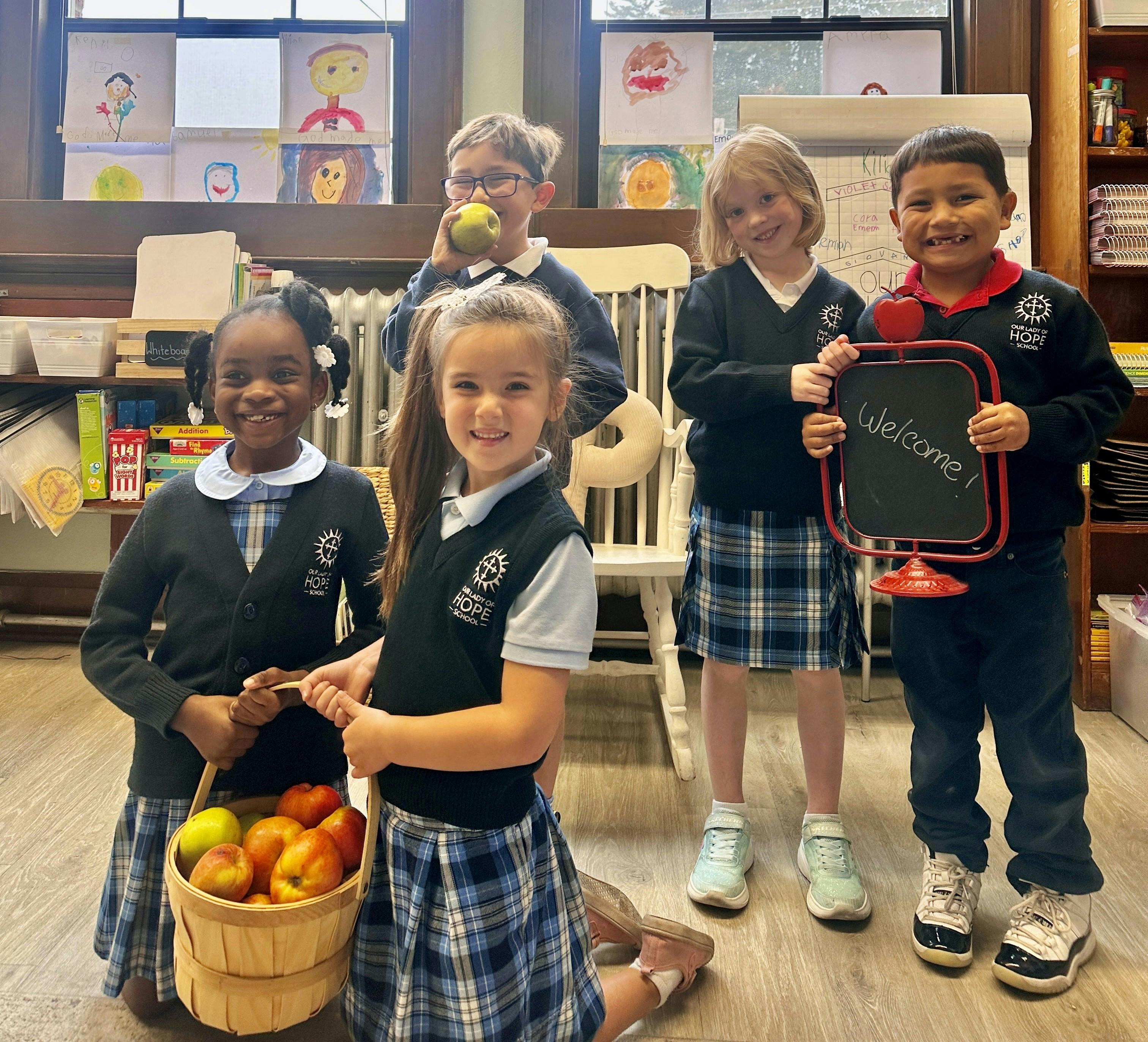 Five children in uniform holding a basket of fruit and a small chalkboard with "Welcome"