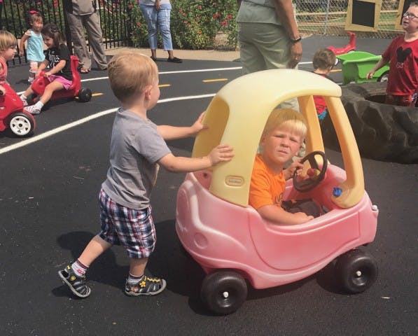 Child pushing another child in a toy car on a paved surface