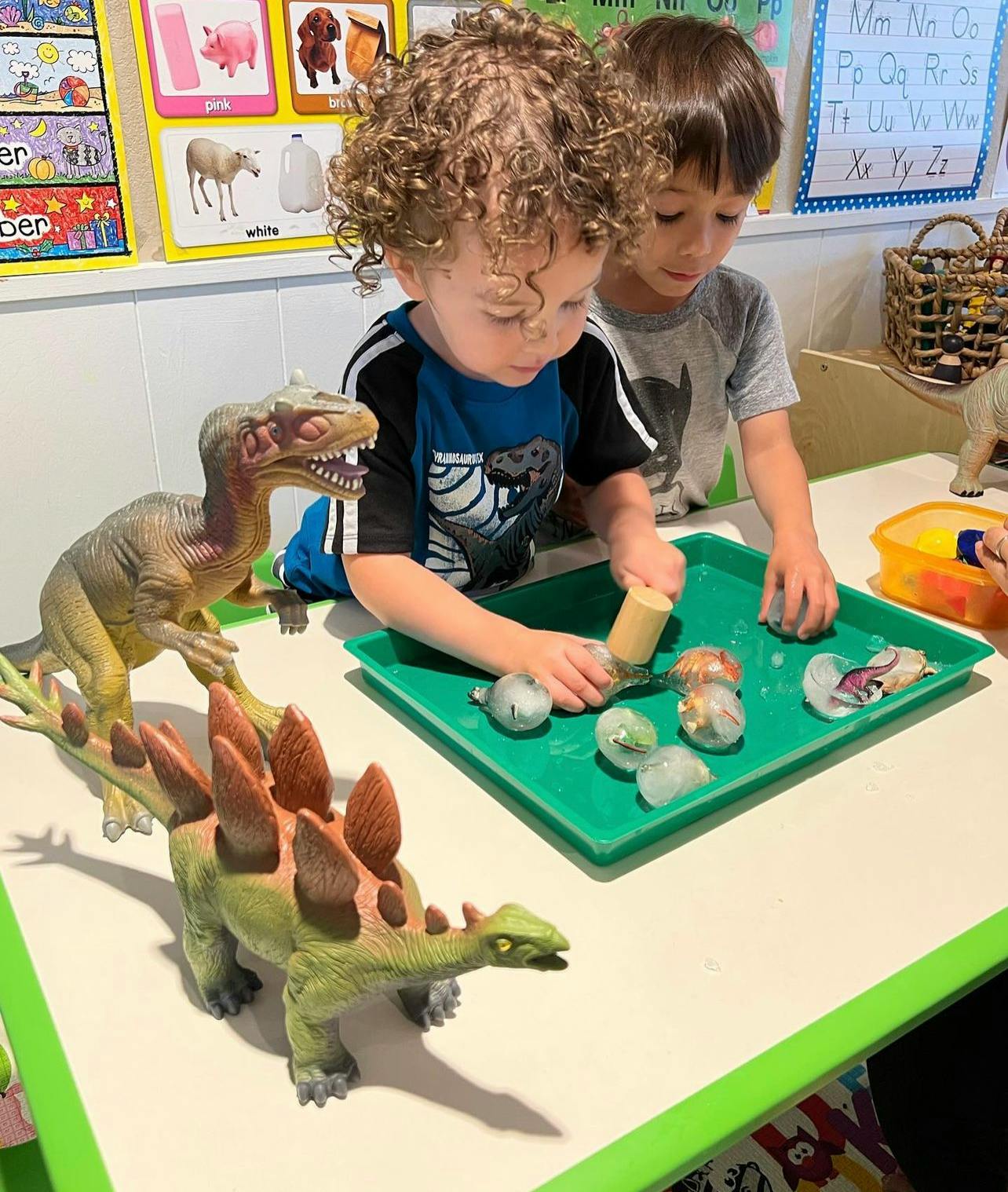 Two children playing with dinosaur toys and a green tray with small objects