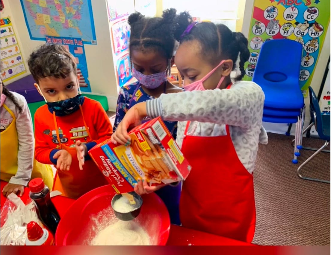 Children in masks pouring ingredients into a bowl in a classroom
