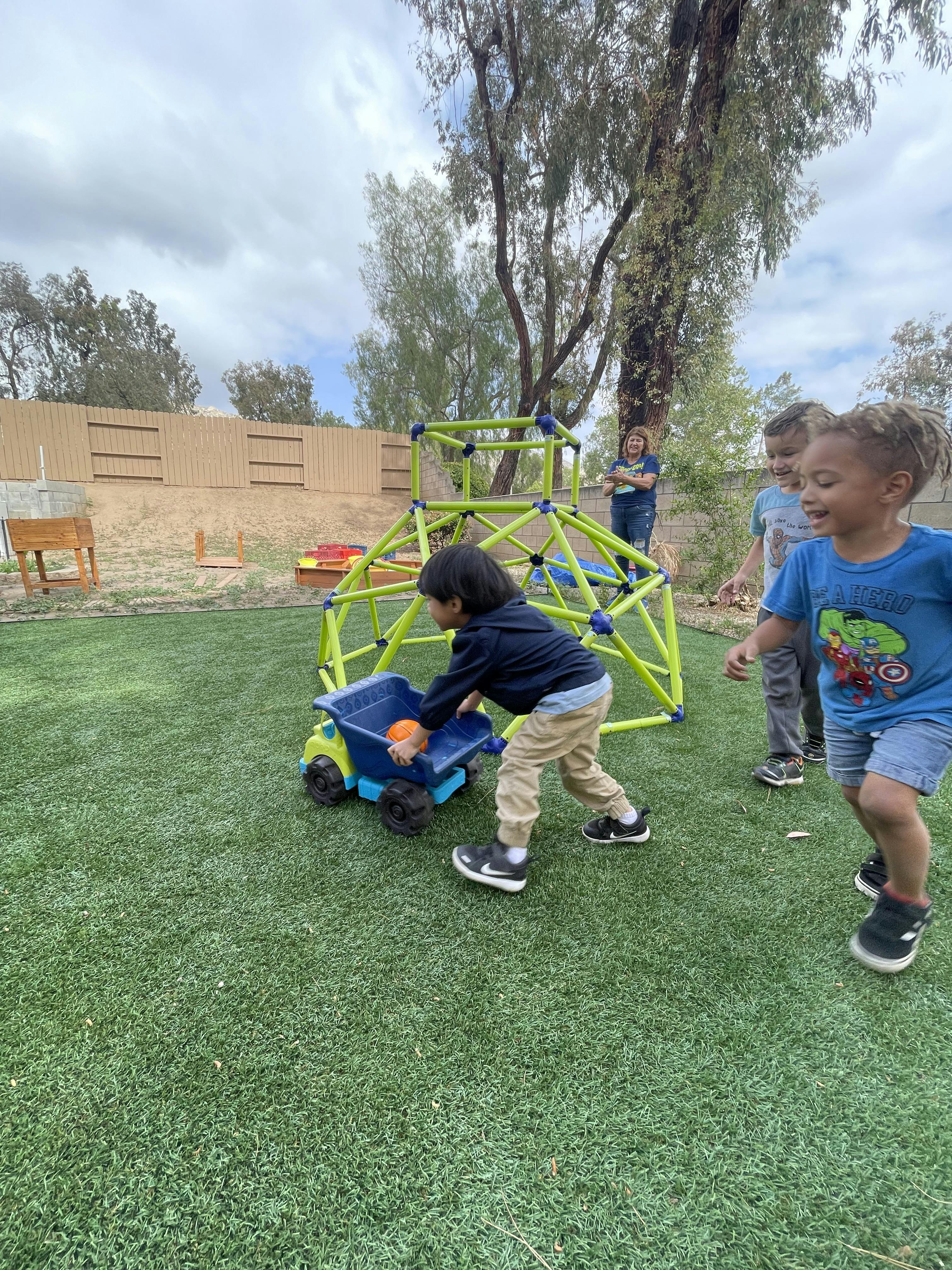 A child pushing a toy truck with an orange ball in the back on green turf, two other children are to the right of them on the turf, and a person is standing in the background
