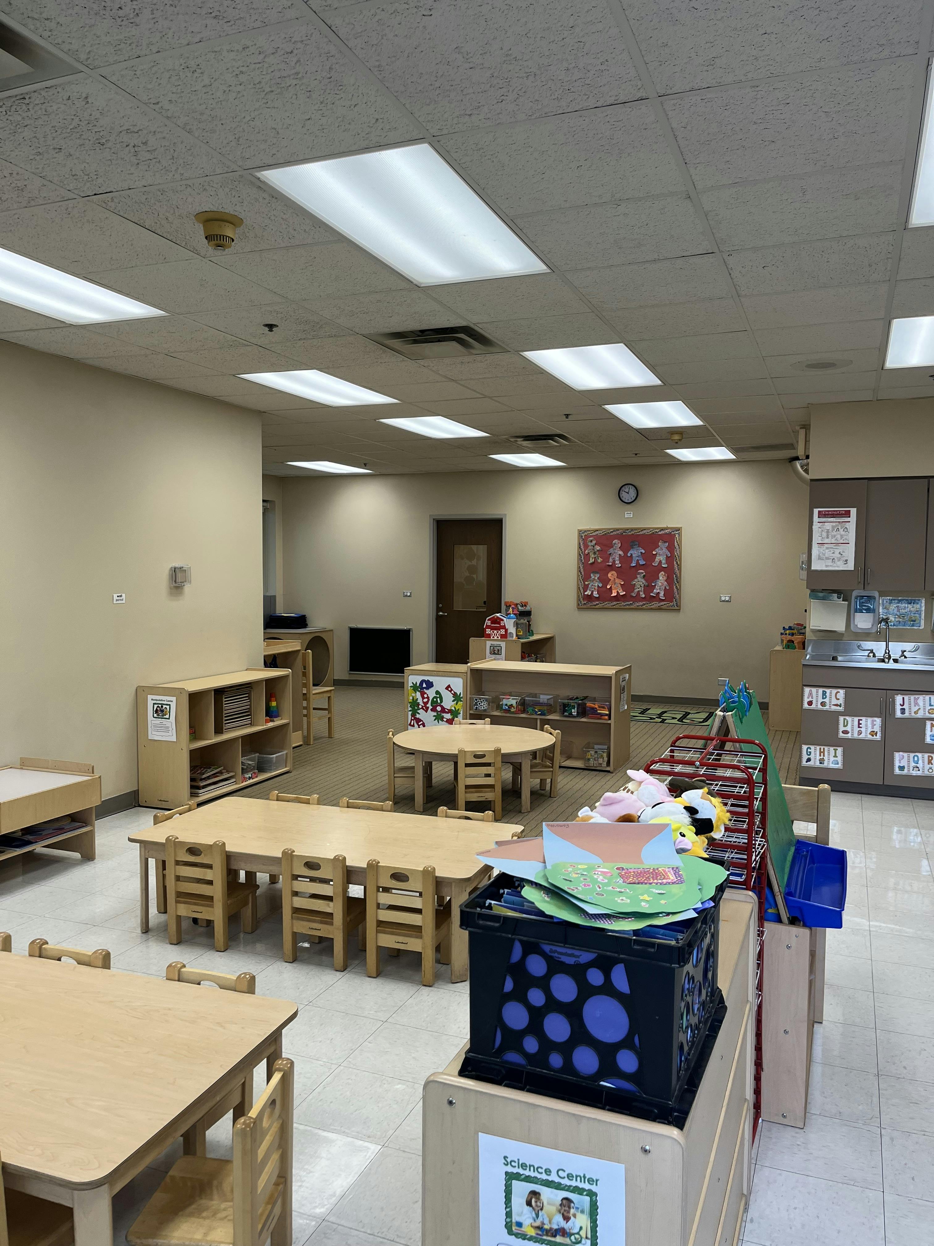 Empty classroom with small wooden tables and chairs, shelves with toys