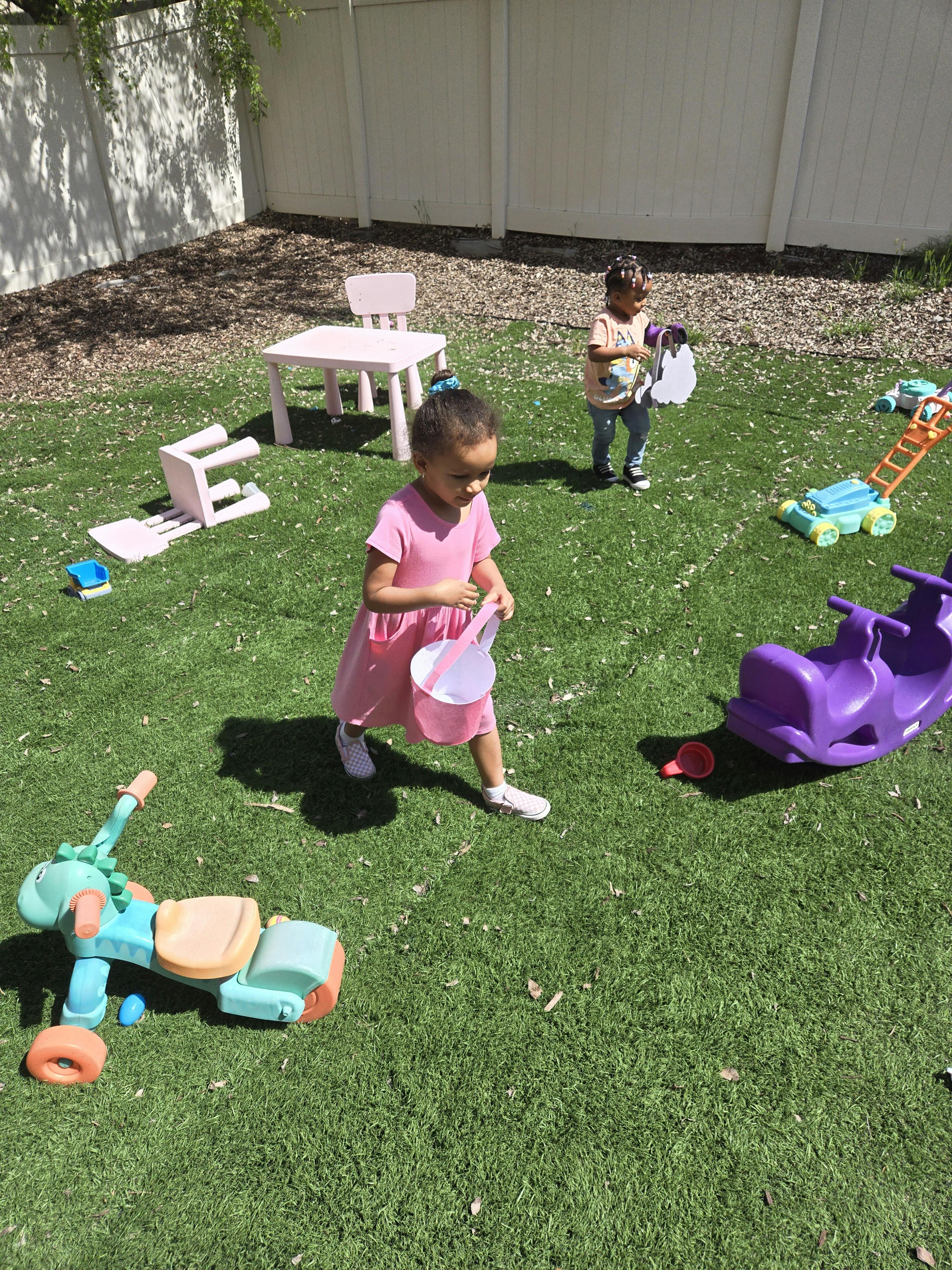 Child in pink dress walking with a bucket in a grassy yard with toys