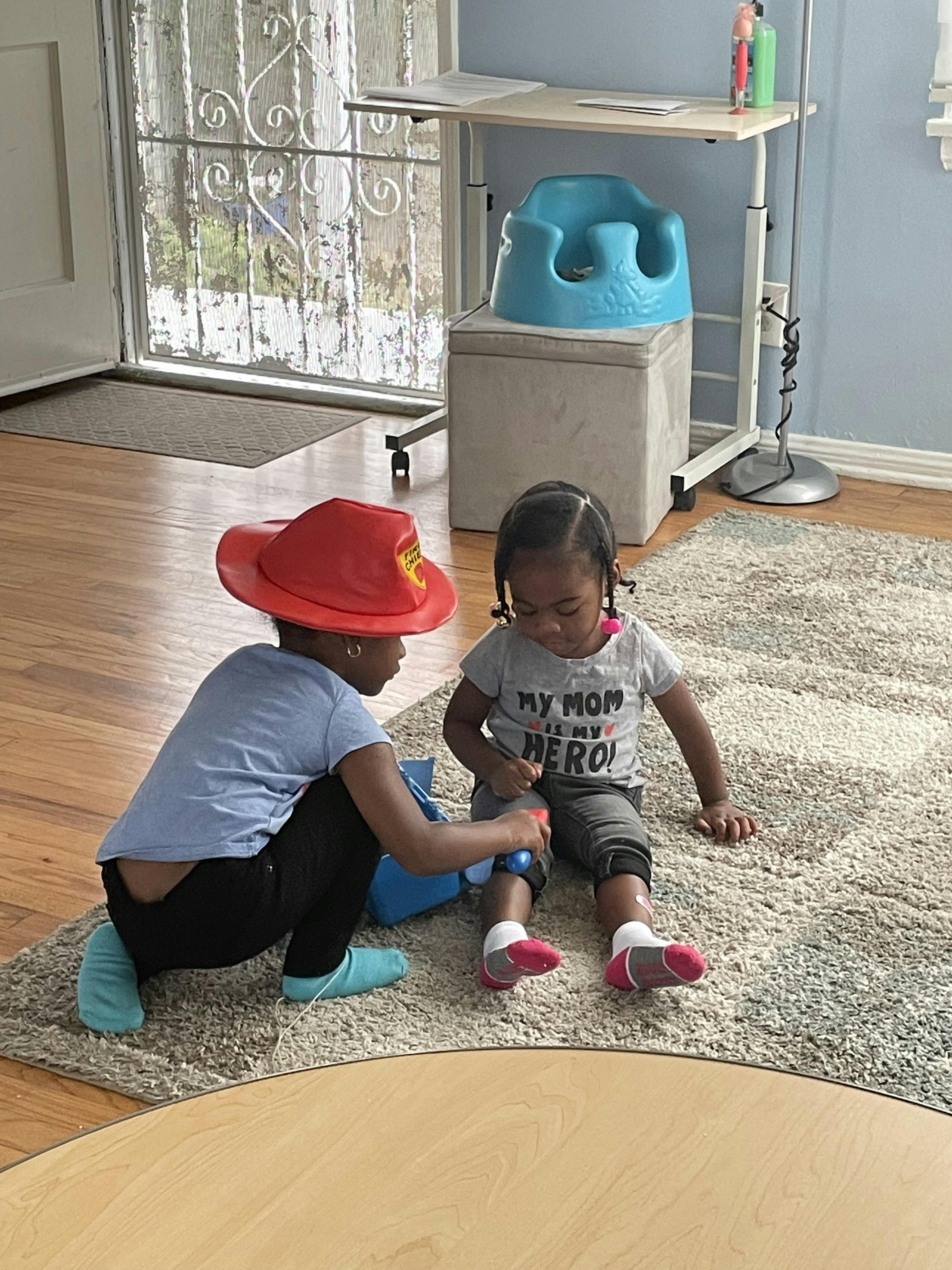 Child in red hat pretending to spray a toy bottle on another child's knee