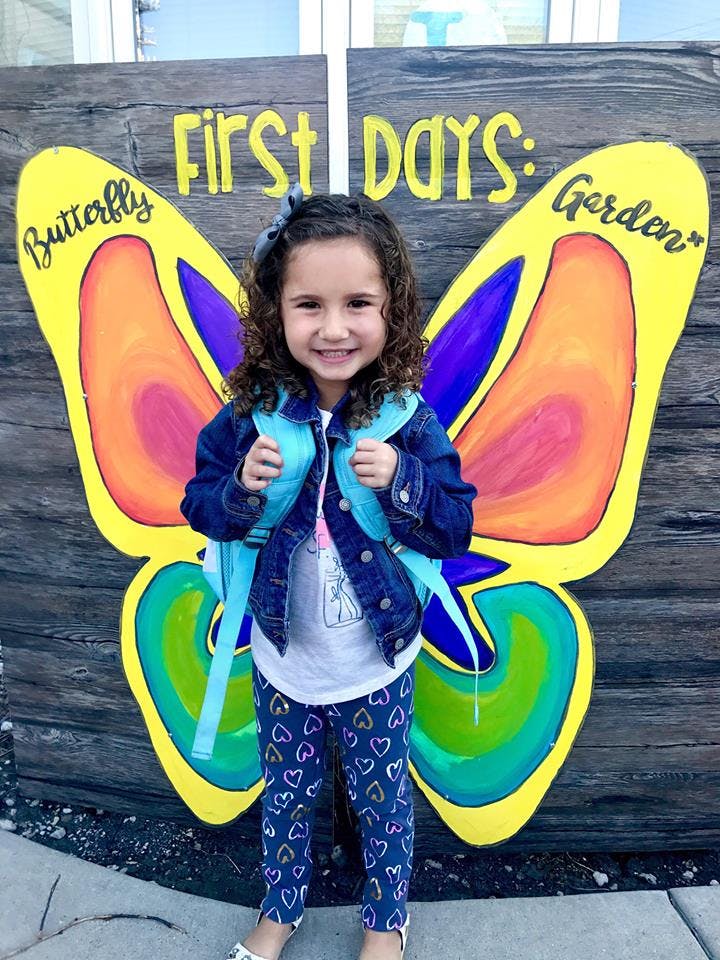 Child wearing a backpack standing in front of a butterfly cutout with text "First Days"