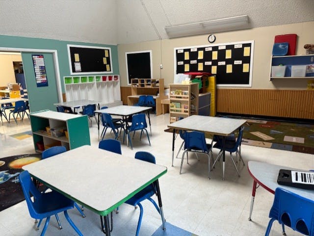 Empty classroom with small tables and chairs arranged, a bulletin board, and shelves