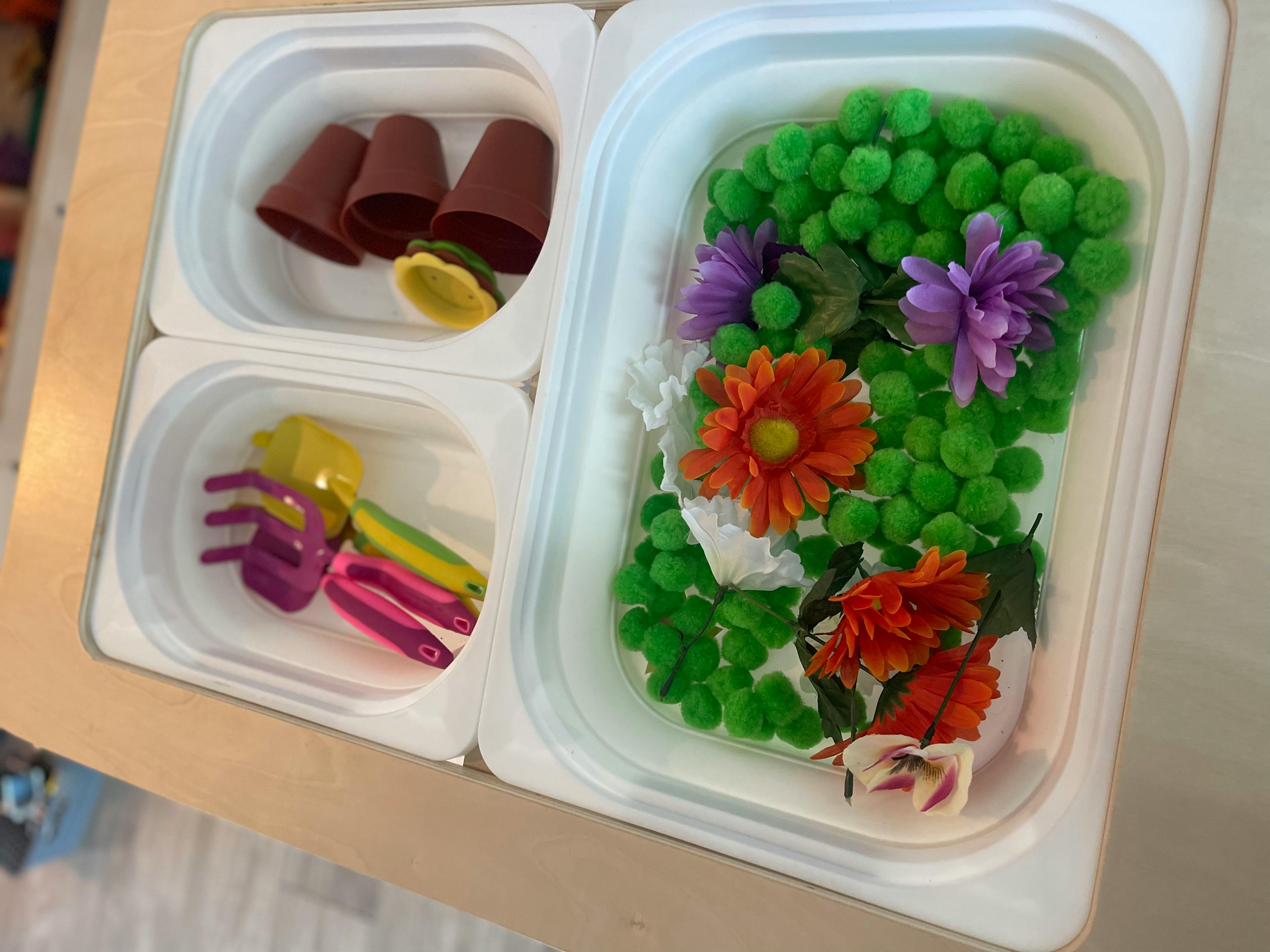 Sensory bin with gardening pots, tools, green pom-poms, and artificial flowers