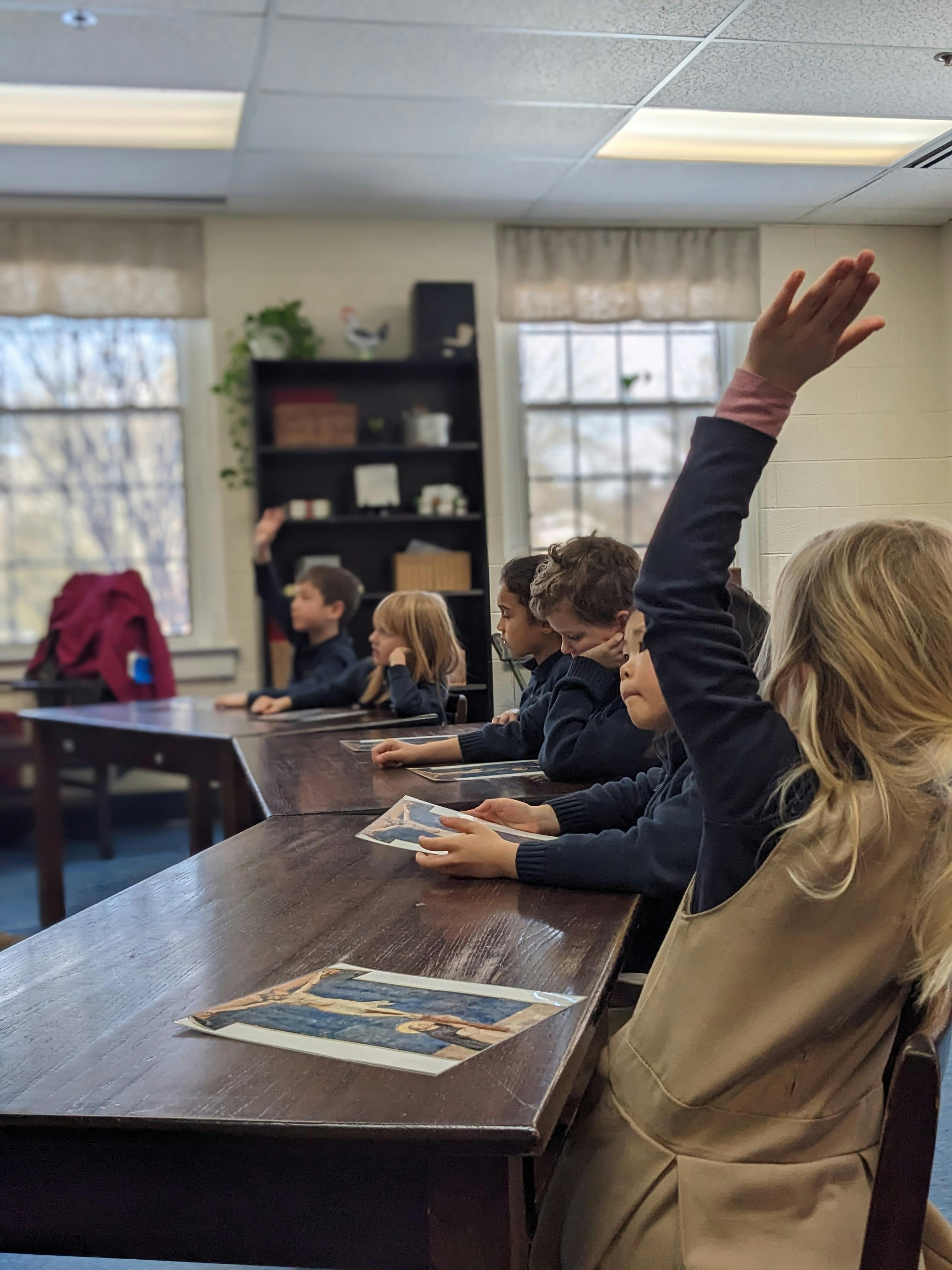 Children sitting at desks in a classroom, one with a hand raised