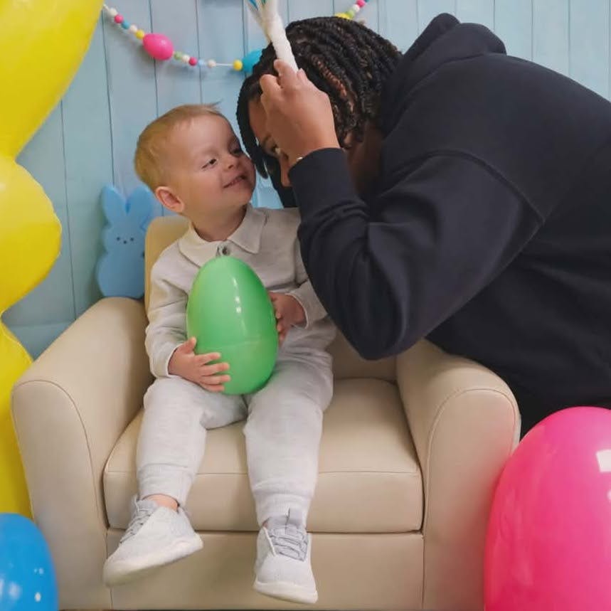 A person placing bunny ears on a child sitting in a chair holding an egg